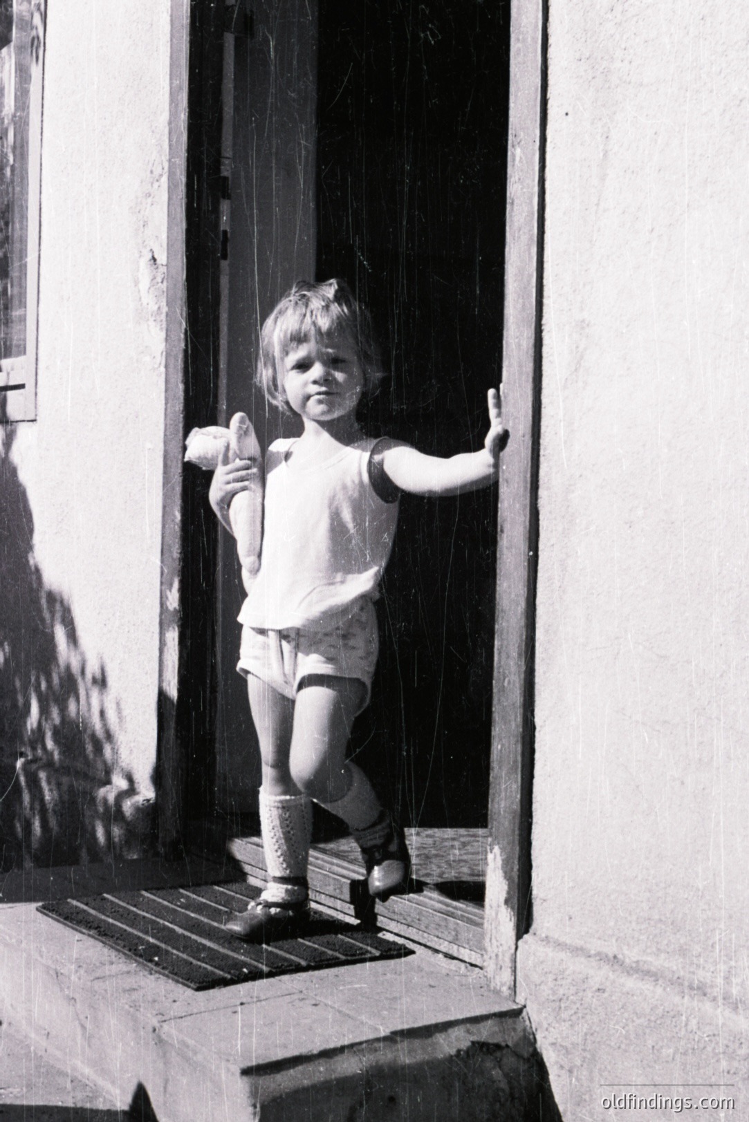 A young child stands in a doorway, holding an object (possibly bread) in their hand. The child wears a simple sleeveless top, short shorts, socks, and shoes. Architectural details suggest a modest, older structure. Likely a candid family snapshot, possibly 1960s-1970s. Evokes nostalgic, domestic scenes.