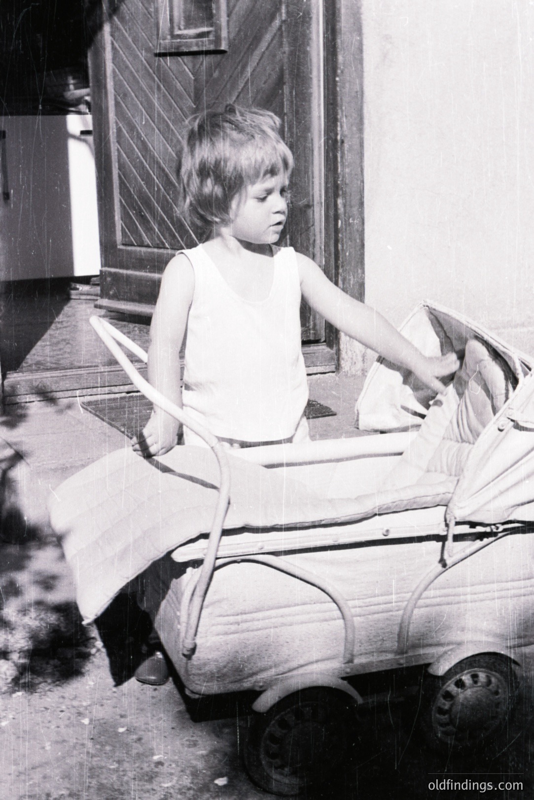 A young boy sits in a metal toy car resembling a vintage European automobile. He wears a simple sleeveless undershirt. Likely 1960s, the image exhibits a candid, documentary style. Black and white print, potentially archival or family snapshot. High commercial value for nostalgic design references.