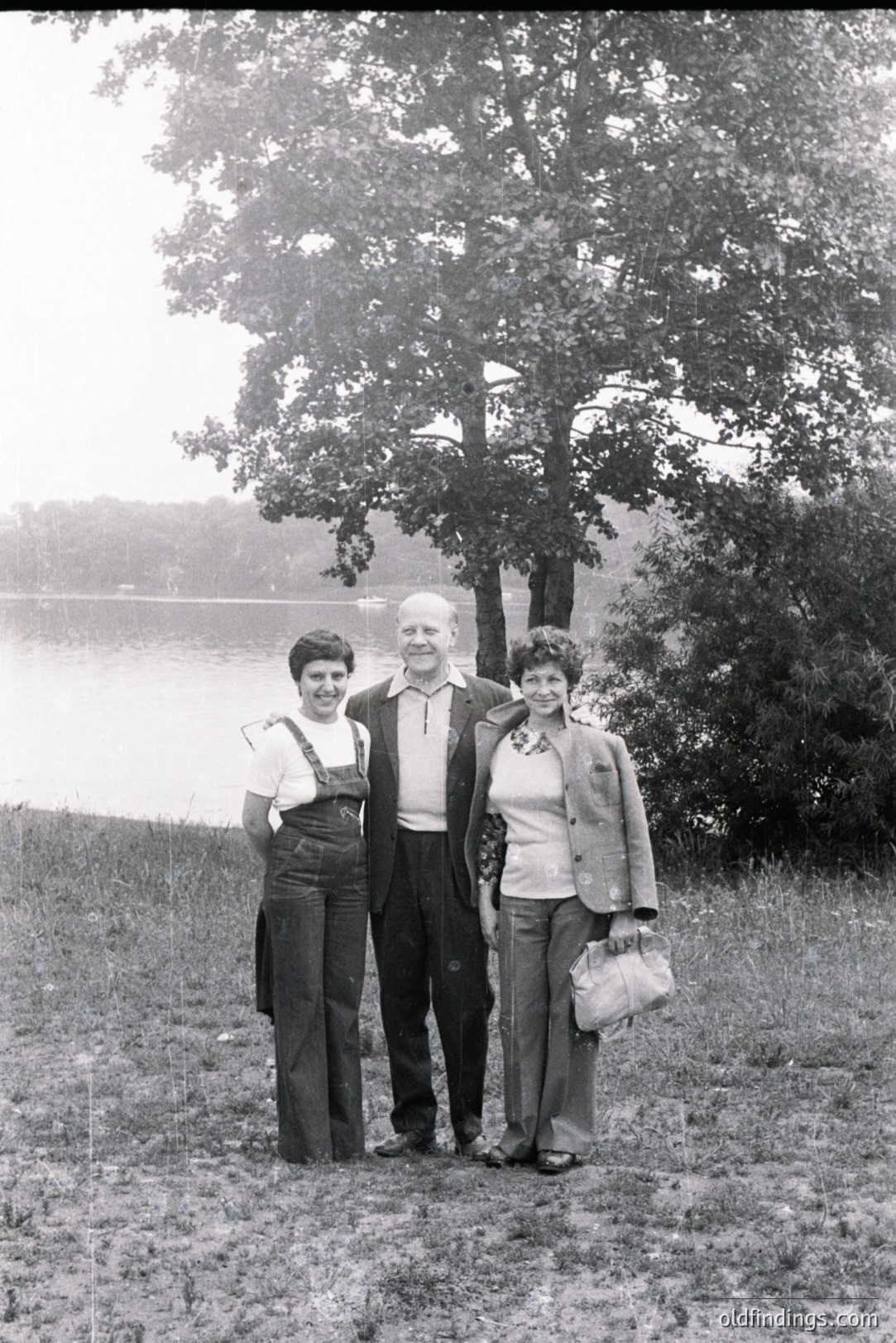 Three individuals stand lakeside, likely a family portrait. The man wears a suit jacket; the women, bell-bottom jeans and casual tops with outerwear. The scene suggests a relaxed, outdoorsy lifestyle, possibly a vacation. Appears to be 1970s style.