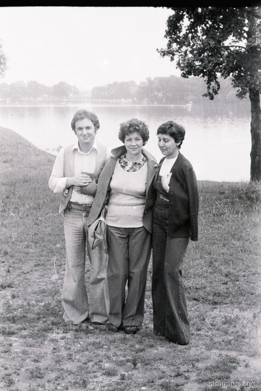 A family portrait from the 1970s: three individuals stand on a grassy bank overlooking a lake. The man wears a patterned vest, flared trousers, and a collared shirt. The woman has a statement necklace and a dark coat, while the younger person wears a dark turtleneck and flared jeans.