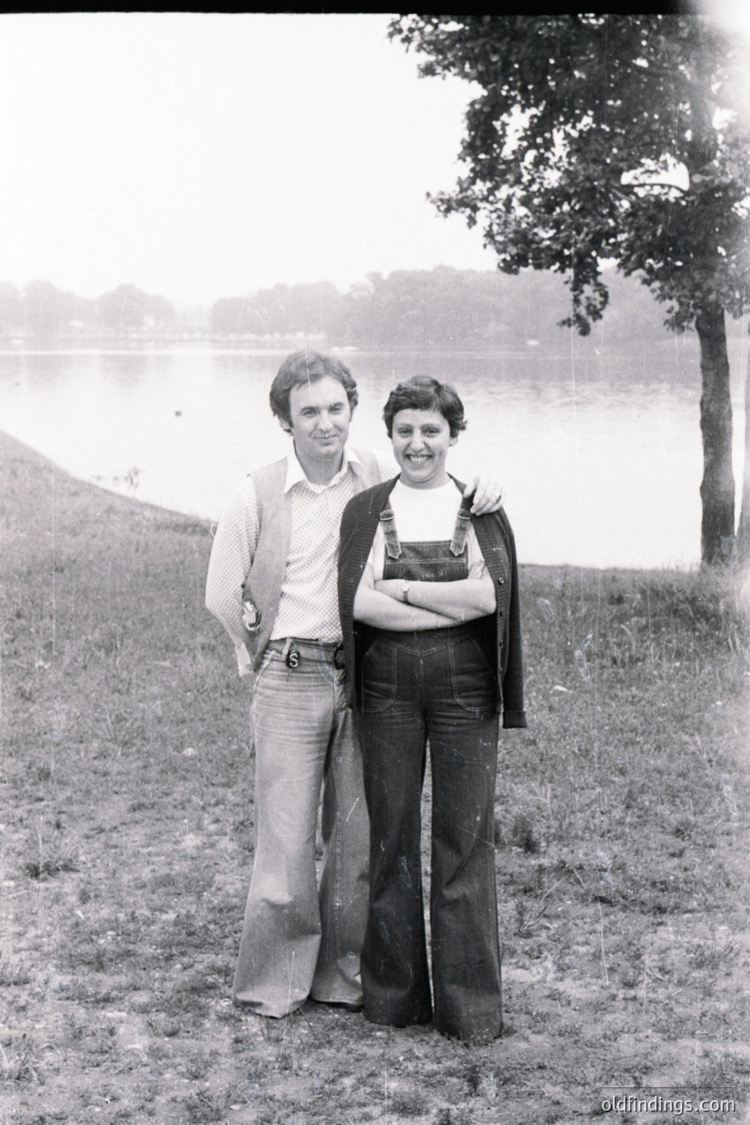 A couple stands on a grassy hill overlooking a body of water. Man in flared trousers, a button-down shirt, and a cardigan. Woman in bell-bottom jeans, a dark turtleneck, and a cardigan. Likely mid-1970s fashion. A lake and trees visible in the background. Candid moment; a casual portrait.