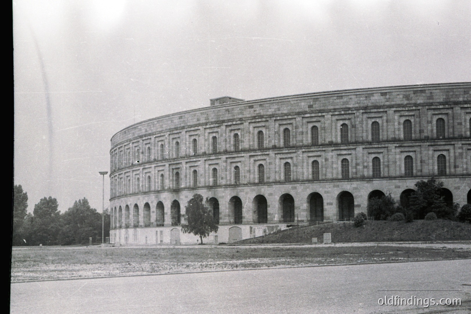 Architectural study of a monumental, curved structure with repetitive arched windows, possibly a gallery or promenade. Formal landscaping with clipped hedges and trees is visible below. Likely a government or institutional building, potentially 1960s-1970s.