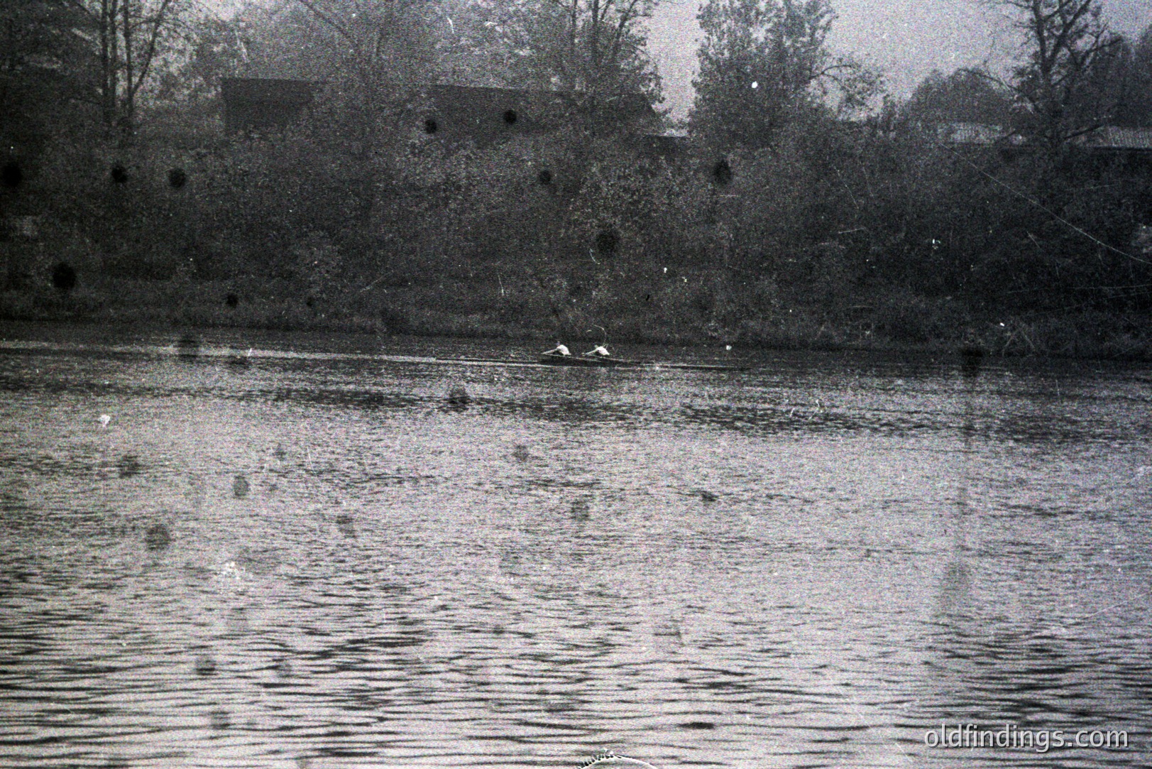 A pair of rowers in a small boat navigate a rippled body of water. The background features indistinct trees and a building, possibly industrial, obscured by mist or distance. Likely 1960s-1970s amateur photography; film grain visible. The overcast lighting and simple composition suggest a documentary or candid moment.