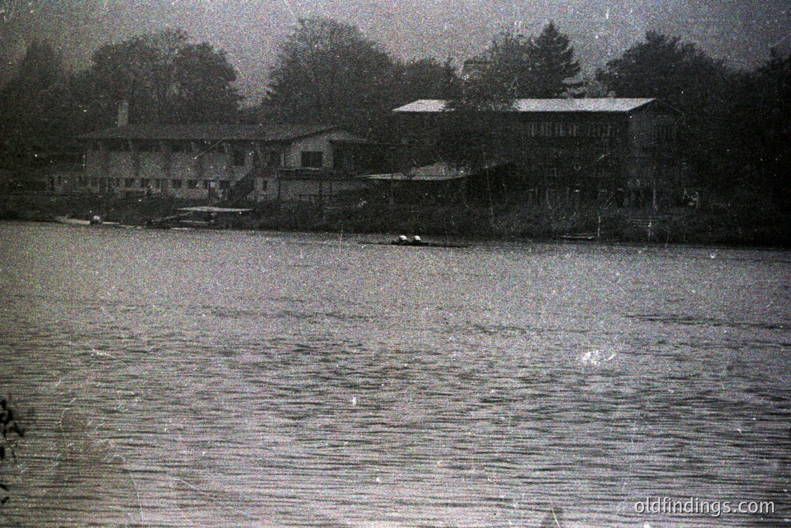 A grayscale image depicts a waterfront scene with a long, low building featuring multiple windows and a gabled roof set back from the water's edge. The structure appears to be a lakeside lodge or resort. Reflections ripple across the water's surface. Trees line the background. Likely 1950s or 1960s, amateur photography.