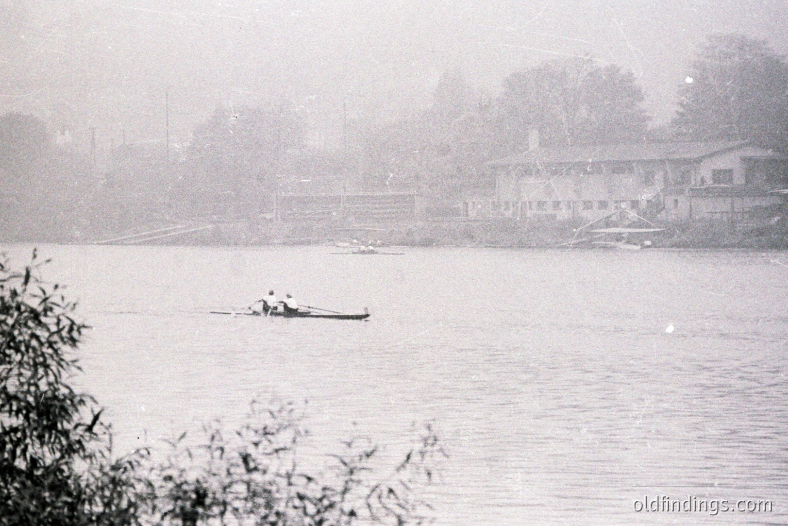 A solitary rower navigates a misty lake, viewed through foreground foliage. A row of lakeside buildings, likely a resort or housing, is visible in the background. Appears to be a vintage amateur photograph, possibly 1960s-70s. Soft focus and grain characterize the image. Likely a local leisure scene.