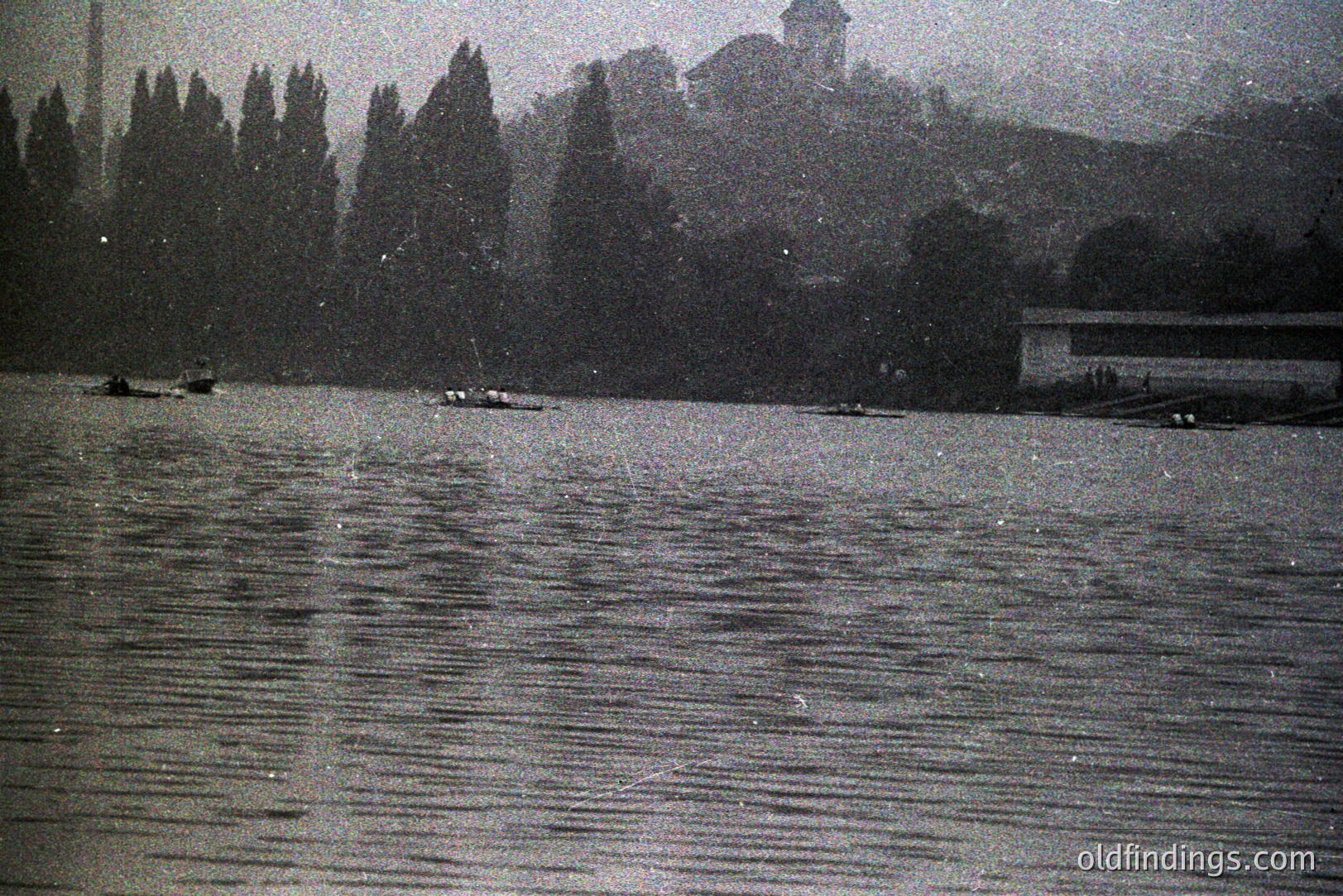 Monochrome image shows rowboats on a body of water, likely a lake or river. Trees line the shore, with a building situated on a distant hillside. Likely a candid snapshot, possibly 1950s-1970s, given the film grain and subject matter. Suitable for vintage aesthetic projects.