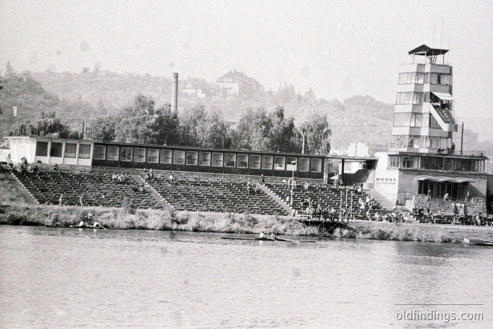 A view of a large, tiered seating structure and a distinctive multi-story observation tower alongside a waterway. Spectators watch rowing events. Likely a regatta or similar sporting venue, possibly from the mid-20th century. Architecture shows modernist influences. Vegetation and hillside backdrop.