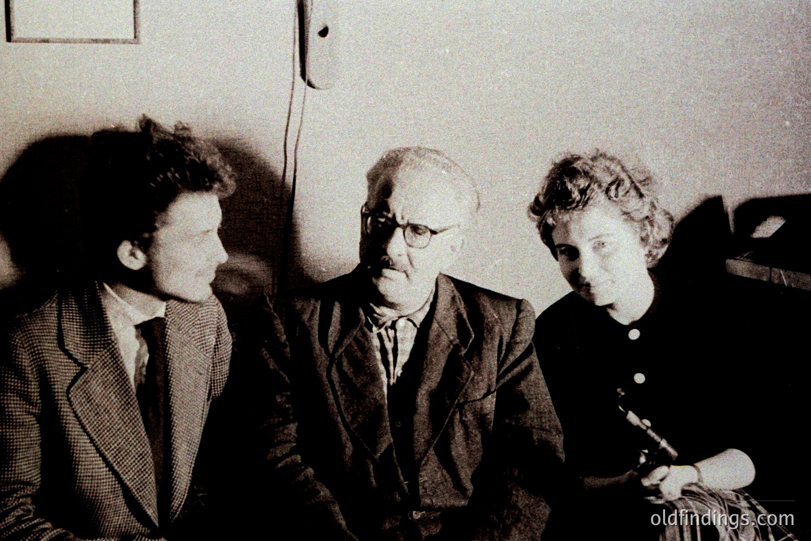 A black and white portrait depicts three individuals in a formal setting. A man with glasses sits centrally, flanked by a young man and a woman holding a microphone. Likely a studio or event, evidenced by backdrop and lighting. Clothing suggests a mid-20th century style (1950s-1970s). Potential commercial value for historical research or design.