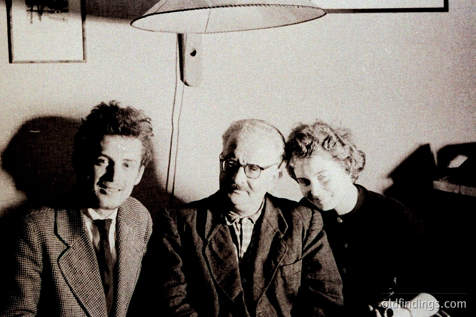 Three men sit closely together, framed by a simple interior. The central figure, bald and wearing glasses, appears more formal in a tailored suit. Flanking him are two younger men with darker, styled hair, also in suits. A lamp casts shadows. Likely a candid portrait.