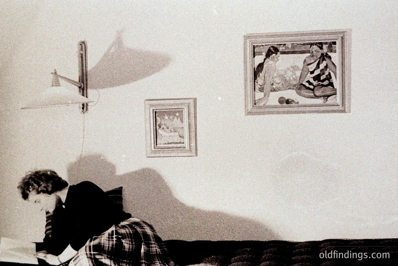 A man sits at a desk, intensely focused on papers, under the light of a wall-mounted lamp. Two framed artworks adorn the wall behind him; one depicting figures, the other smaller and abstract. Likely a study or office scene, 1970s aesthetic.