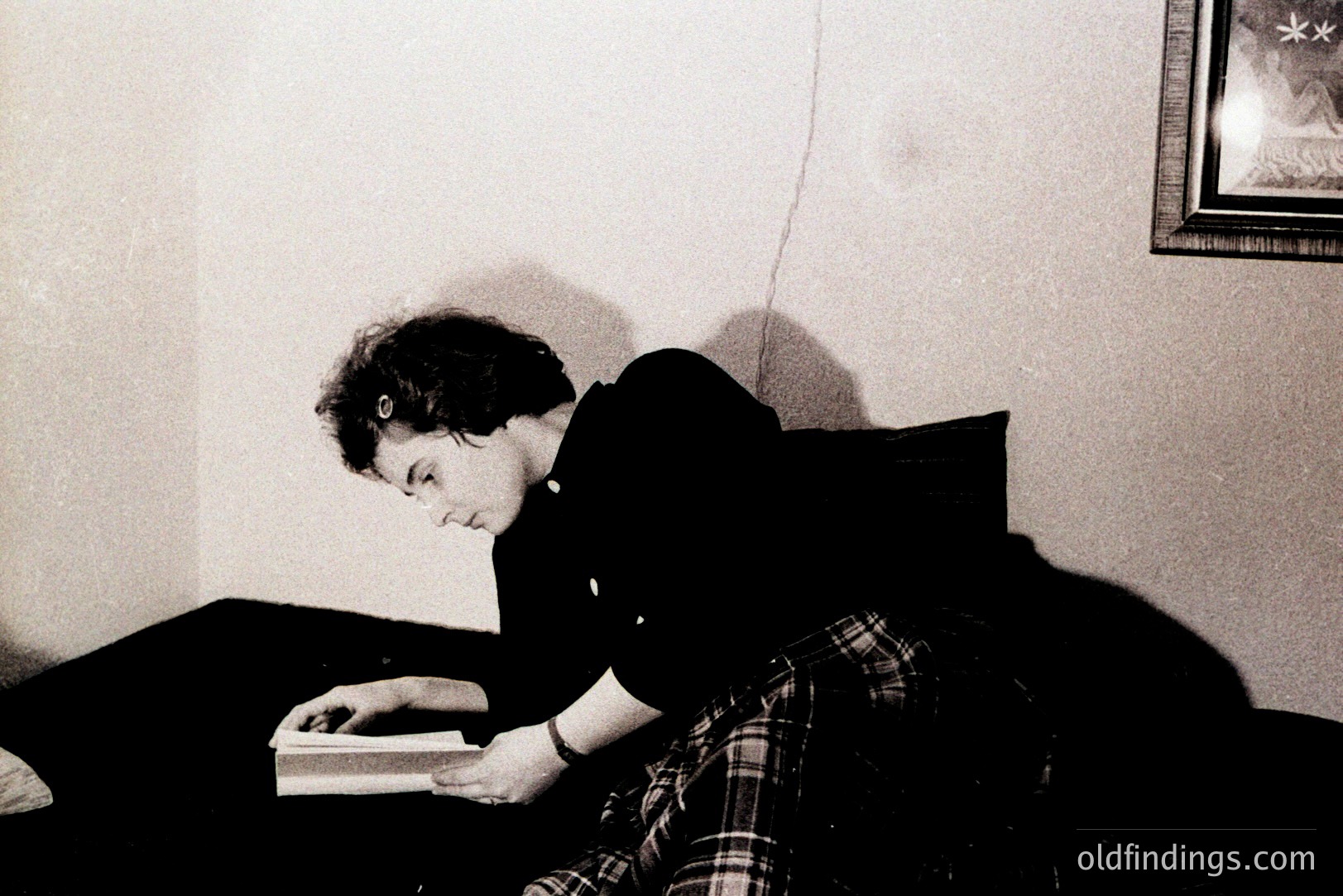 A young woman sits on a cushioned ledge, engrossed in a book. She wears a plaid shirt and black pants. A framed picture hangs on the wall. The composition highlights a relaxed, contemplative atmosphere. Likely a candid shot, circa 1970s-1980s.