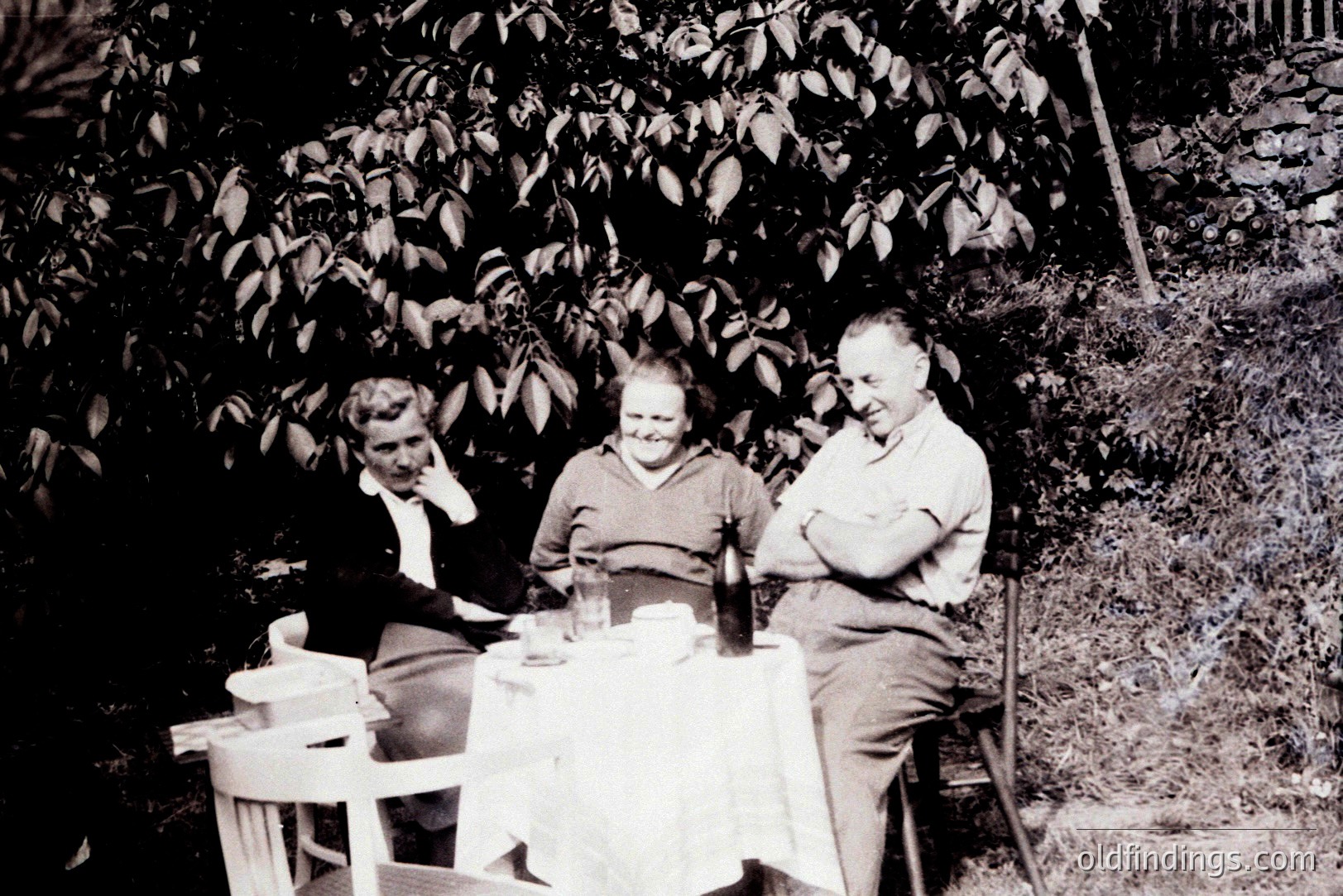 Three individuals seated at a small table covered with a white cloth outdoors. A bottle and glassware suggest a gathering. Dress and hairstyles suggest a 1960s timeframe, possibly a casual, celebratory moment. Natural foliage creates a blurred backdrop. Location is undetermined.