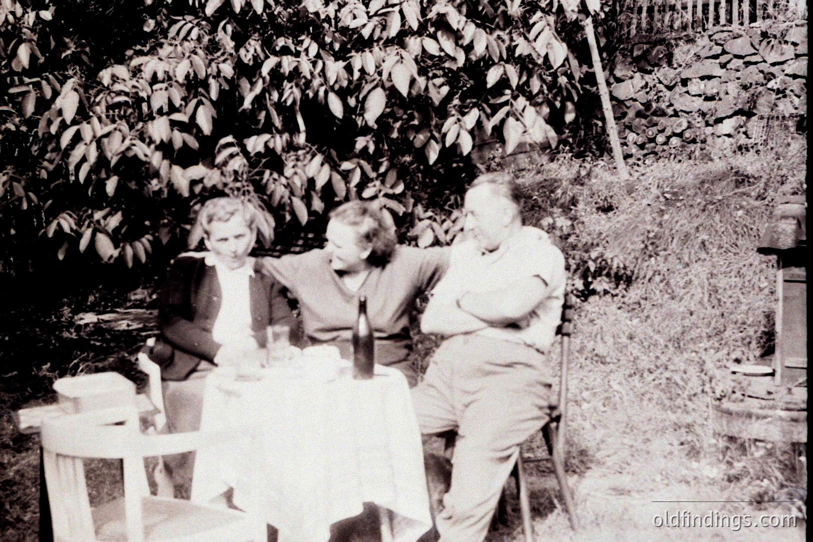 Three figures seated around a table covered with a white cloth, enjoying an outdoor meal. A wine bottle and glassware are present. Lush foliage forms a backdrop. Likely a candid, personal snapshot. Appears to be from the mid-20th century.