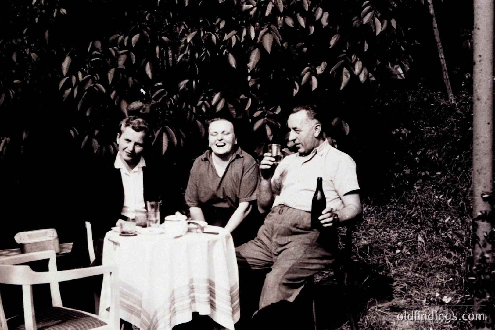 Three people seated around a table laden with drinks, captured in a candid outdoor moment. The scene, likely a garden or patio, is shadowed by foliage. A sense of relaxed leisure and mid-century social gathering is apparent. Likely 1950s-1960s.