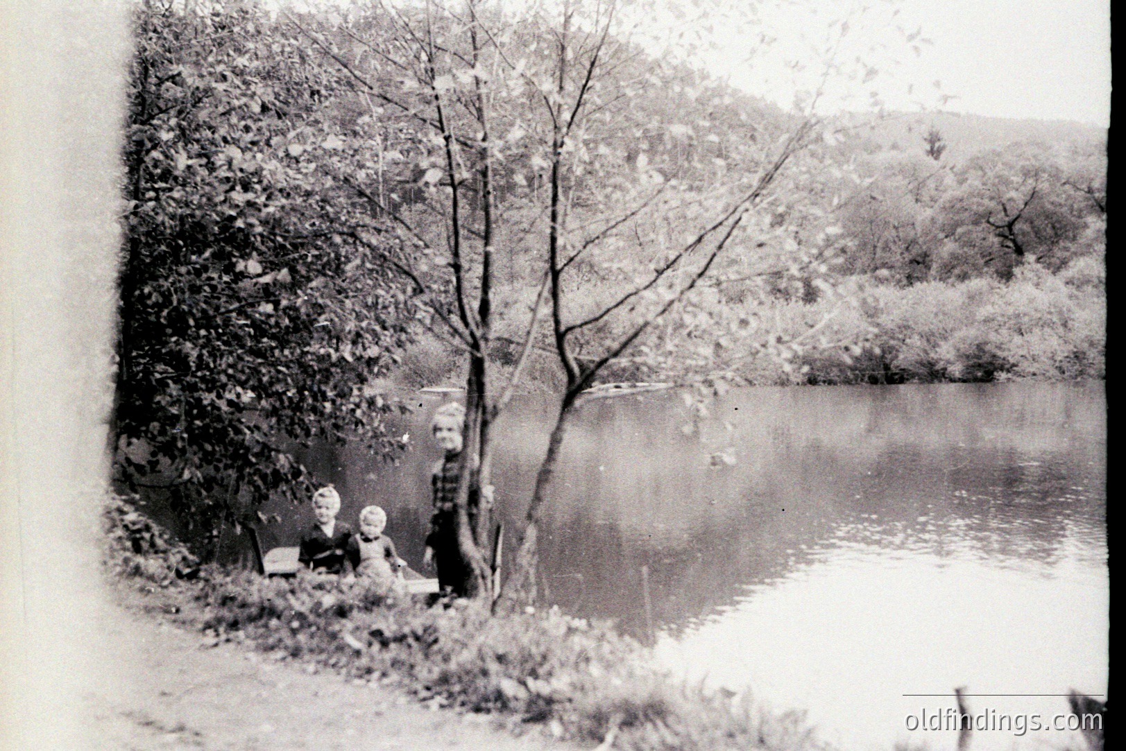 A seated woman and young child pose near a water's edge. Both wear dark, period clothing. A large tree frames the view of the lake beyond. The scene's composition suggests a formal portrait, likely taken outdoors. Style hints at the late 1890s or early 1900s. Location undetermined.