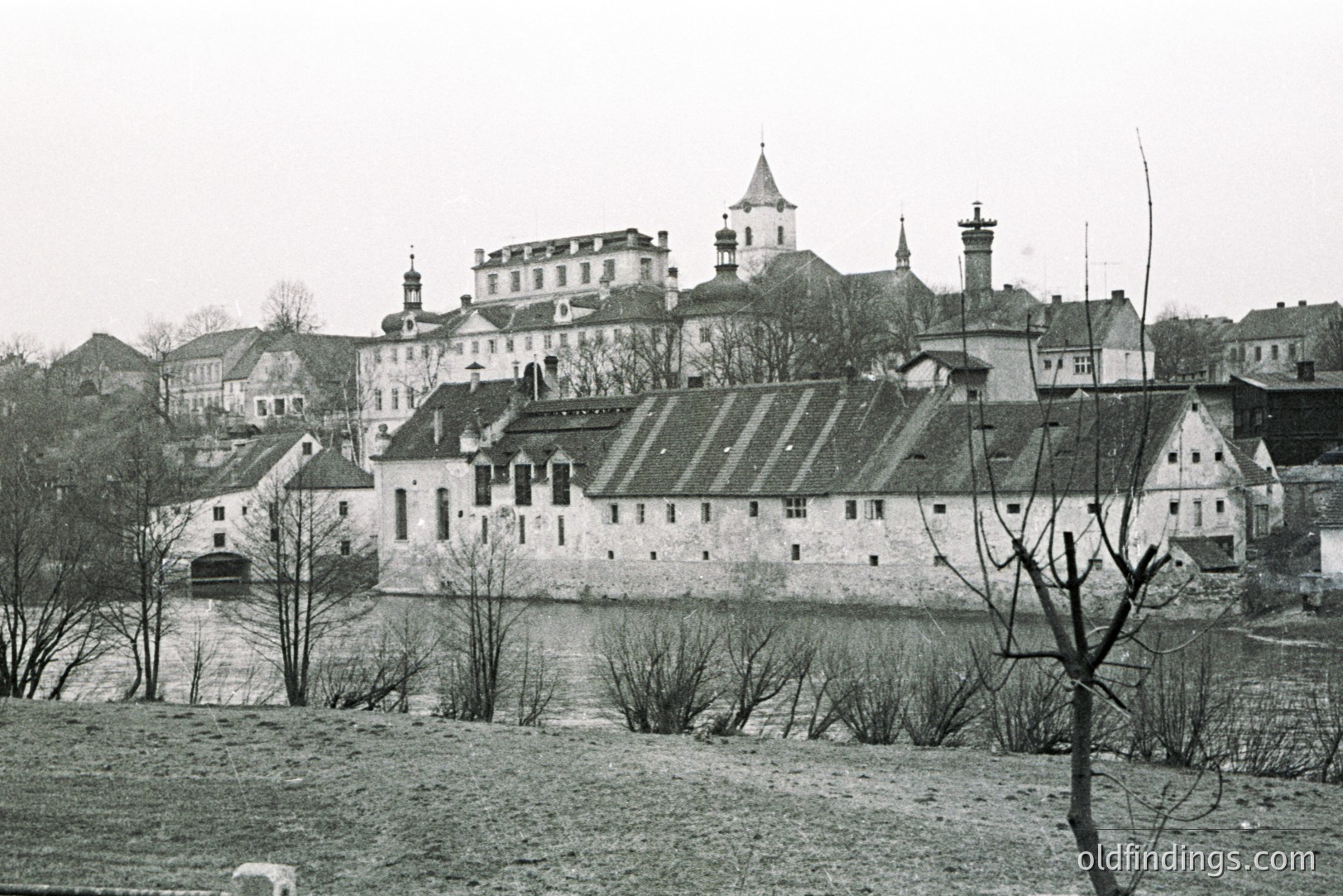 Monochrome view of a fortified structure alongside a river, likely a castle or monastery complex. Prominent white walls and gabled roofs contrast against a gray sky. Buildings rise up a hillside in the background. Likely mid-20th century architectural documentation.