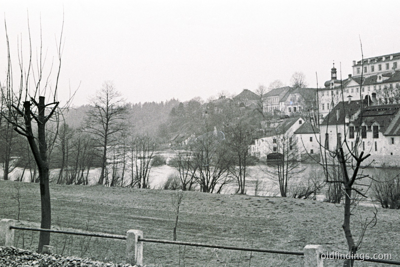 Monochromatic view of a European town, likely Central Europe, featuring a river and a large, ornate building with a steeple. Dense foliage obscures parts of the townscape. Weather appears overcast, possibly snowy. A stone wall and bare trees dominate the foreground. A typical aesthetic of early to mid-20th century travel photography.