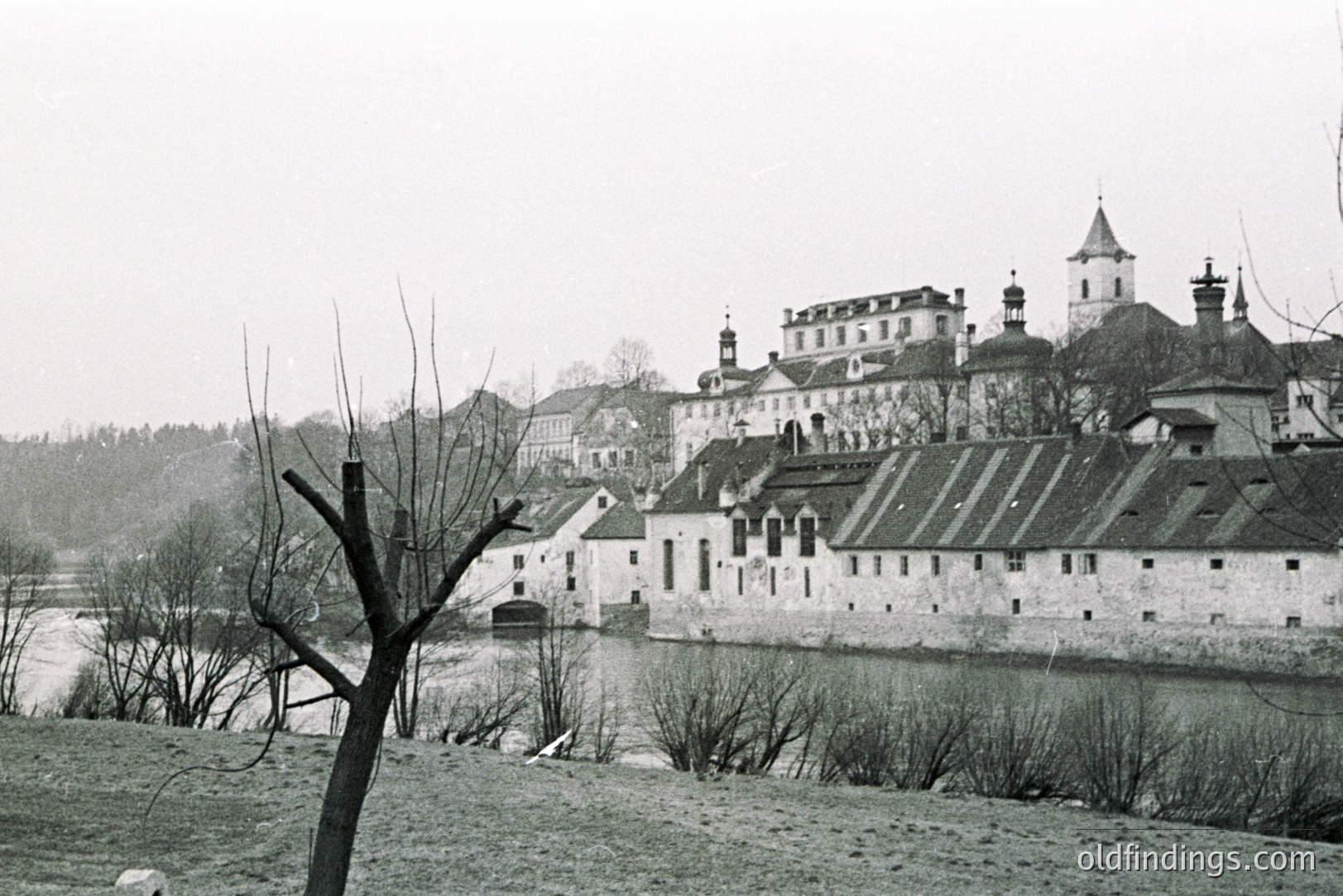 Overlooking a river, a stately castle and adjacent structures dominate the landscape. A stone wall runs parallel to the waterway, with various buildings exhibiting gabled roofs and ornate detailing. The castle features a central tower and multiple chimneys. Likely Central European, mid-20th century.