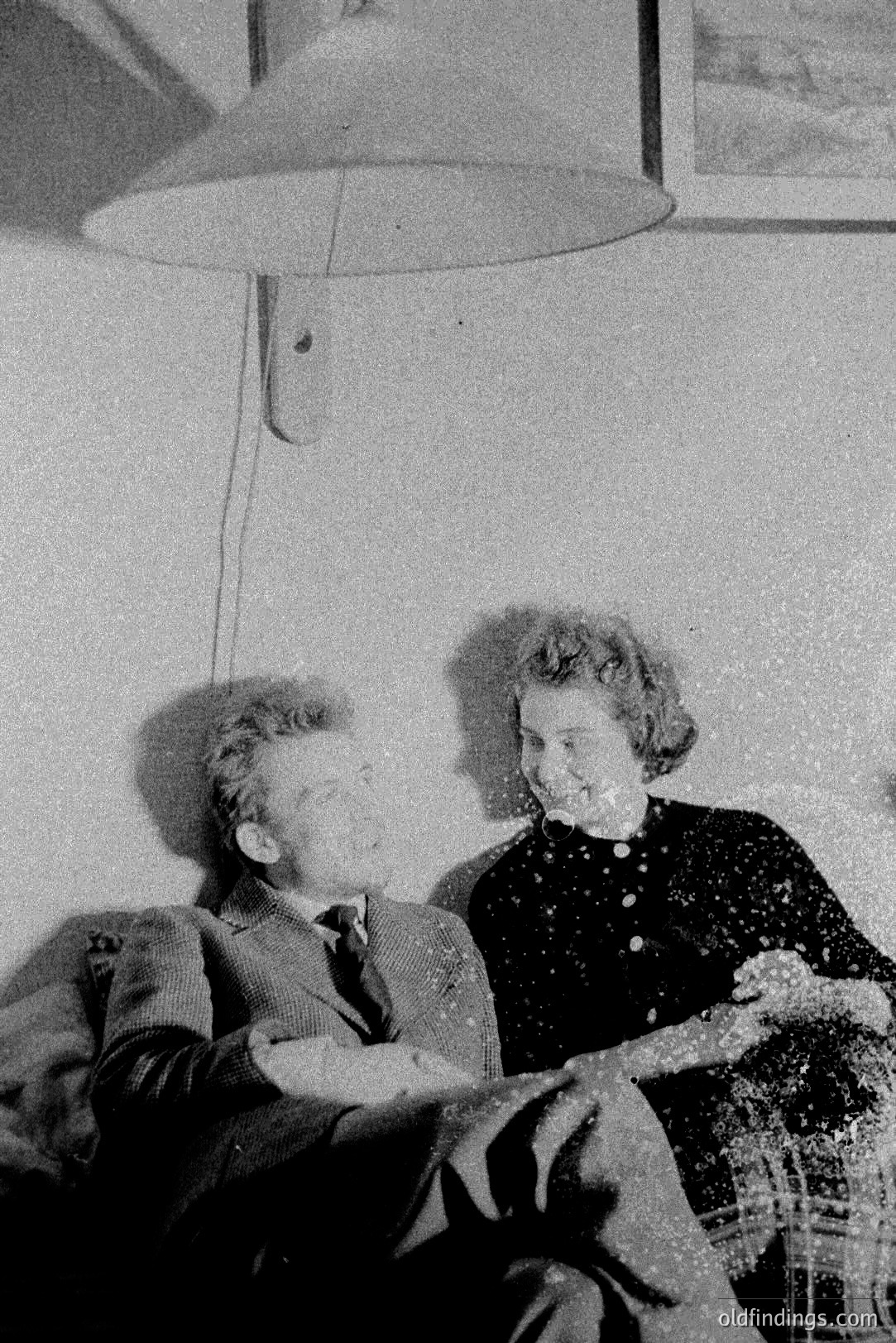A candid, black and white shot captures a man and woman seated and laughing. The man, in a suit and tie, sits on a cushioned chair, while the woman, wearing a dark dress, playfully tosses what appears to be flour or powder into the air. A modern pendant lamp hangs overhead. Likely 1960s.