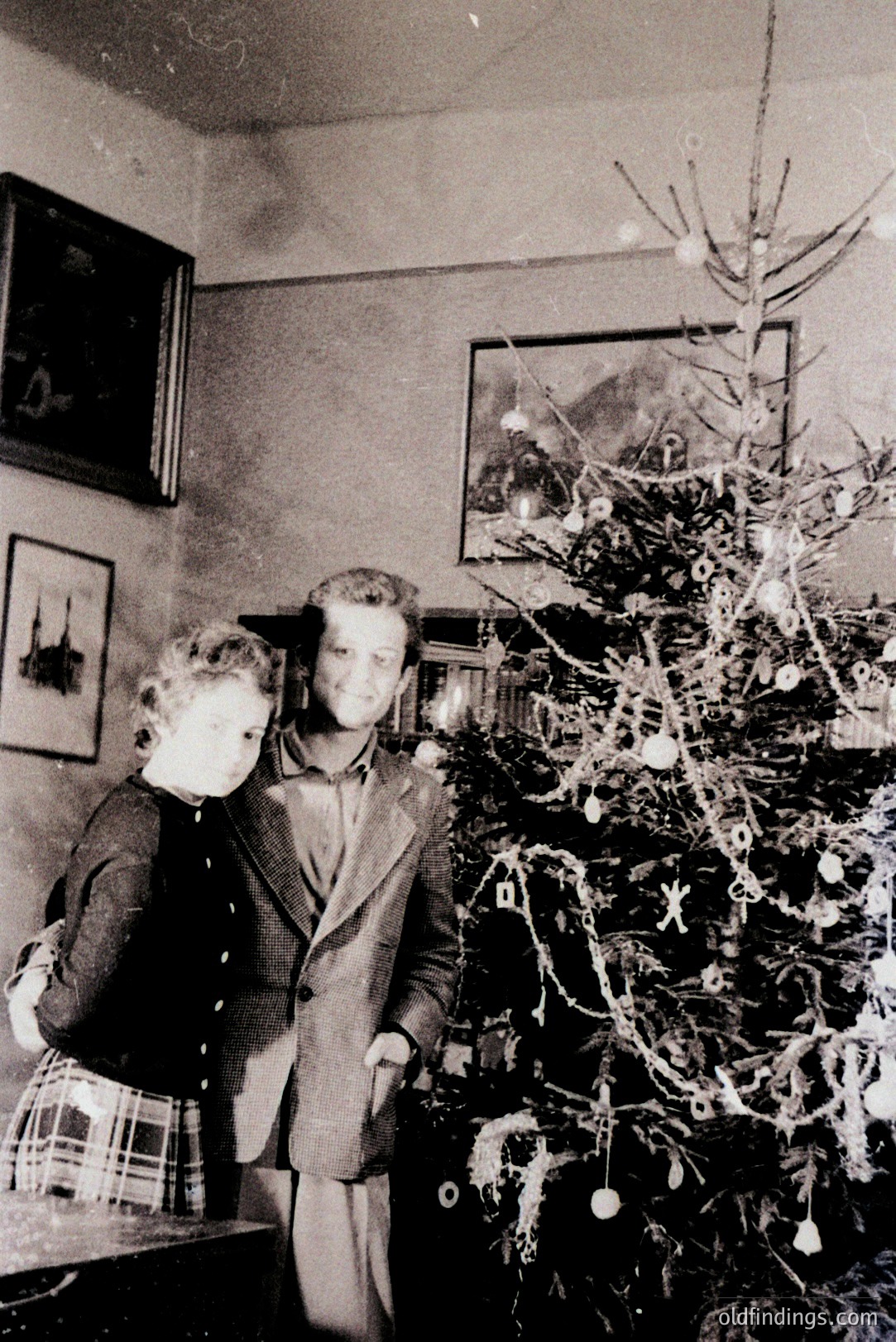 A young couple stands beside a sparsely decorated Christmas tree in a dimly lit room. The tree displays simple ornaments and likely dates to the mid-20th century. Framed artwork adorns the walls, suggesting a middle-class home. A plaid skirt is visible on the woman. 1950s family portrait.