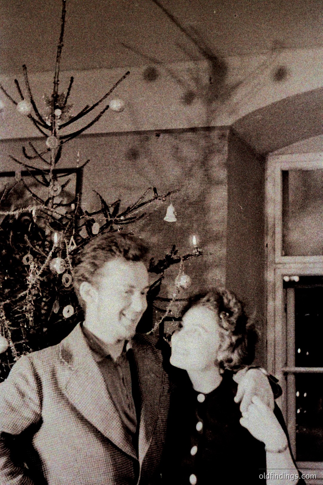 A couple embraces in front of a sparsely decorated Christmas tree, captured in a candid moment. The room features a visible beam structure and a window to the side. Likely 1940s-1950s, showcasing mid-century domestic life. A charming, nostalgic portrait.