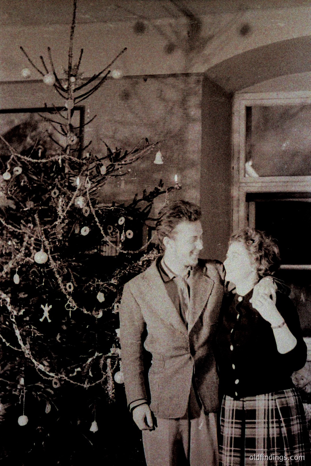 A formal portrait captures a young couple standing beside a sparsely decorated Christmas tree, likely 1950s. The man wears a suit and tie, while the woman dons a plaid skirt and a stylish, short-sleeved top. A window provides natural light. Could be domestic or studio setting.