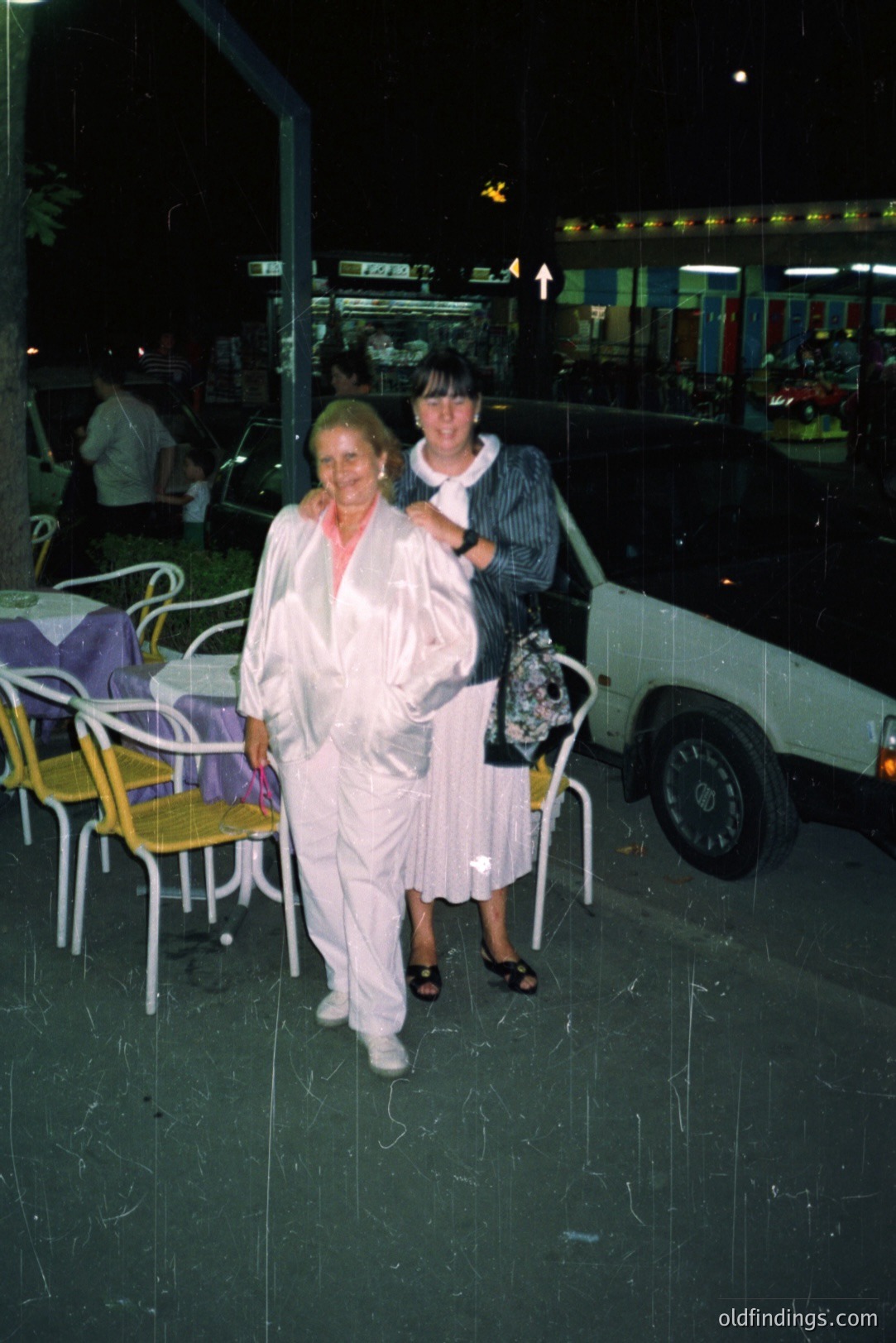 Two women in casual wear pose outdoors at night. One wears a white suit and the other, a floral dress. A restaurant patio with yellow chairs and tables is visible. Likely a seaside resort, possibly Bulgaria. The 1970s-80s aesthetic is apparent. Classic vacation snapshot.
