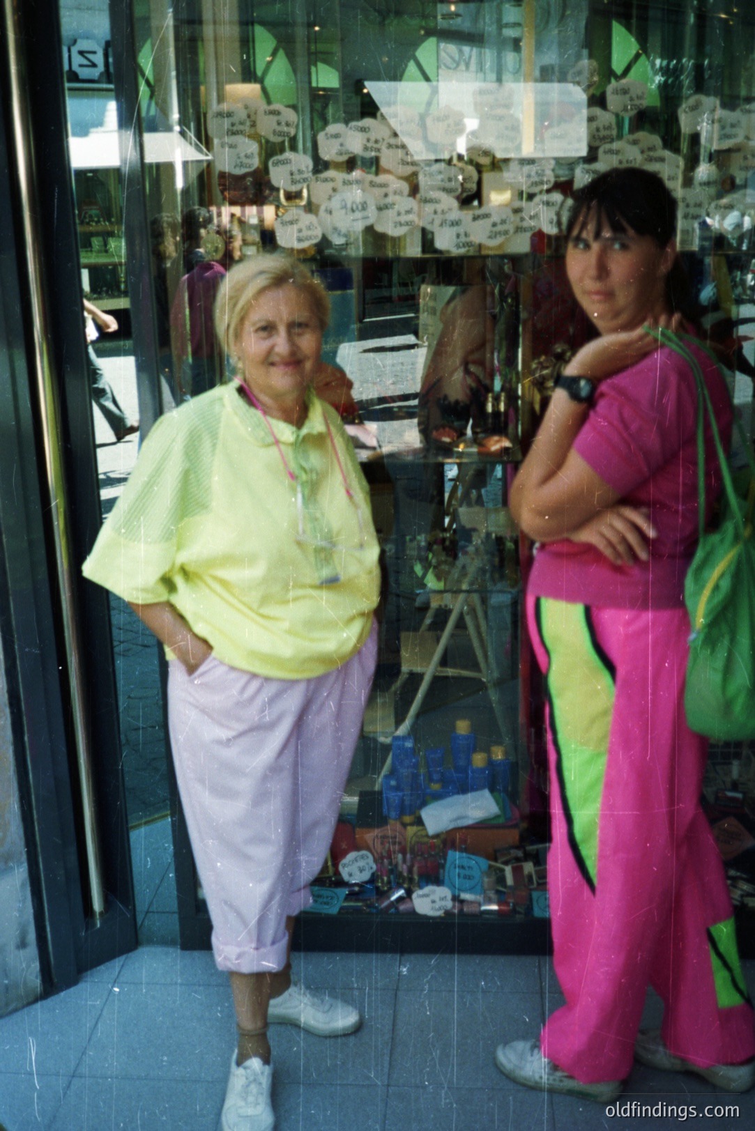 Two women stand outside a store; one in a yellow windbreaker, lilac track pants, and sneakers, the other in a pink and neon green tracksuit and a green bag. The storefront features multiple hanging ornaments. Likely 1980s fashion, possibly travel photography.