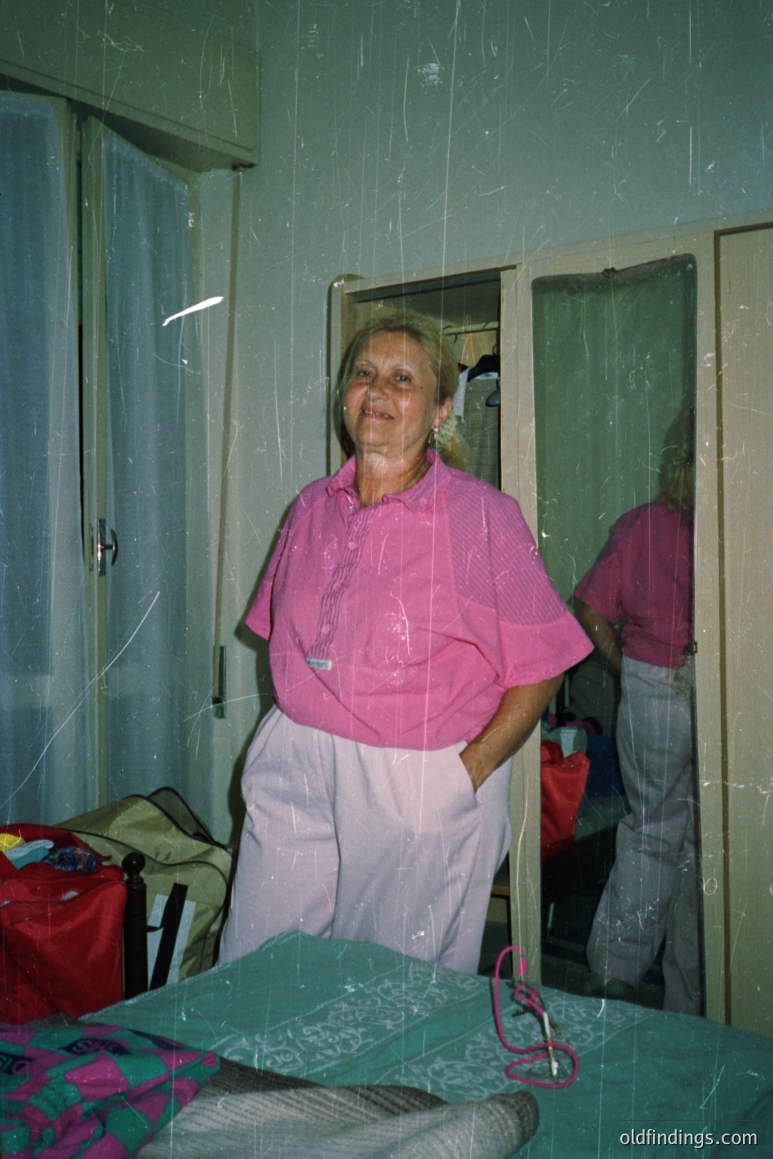 A woman stands in a bedroom, posed casually with hands in pockets. She wears a pink button-down shirt and light-colored pants. The room features a bed, closet with mirror, and minimal décor. Appears to be a snapshot, possibly vacation or family documentation. Likely 1970s-1980s era. Film grain & visible scratches present.