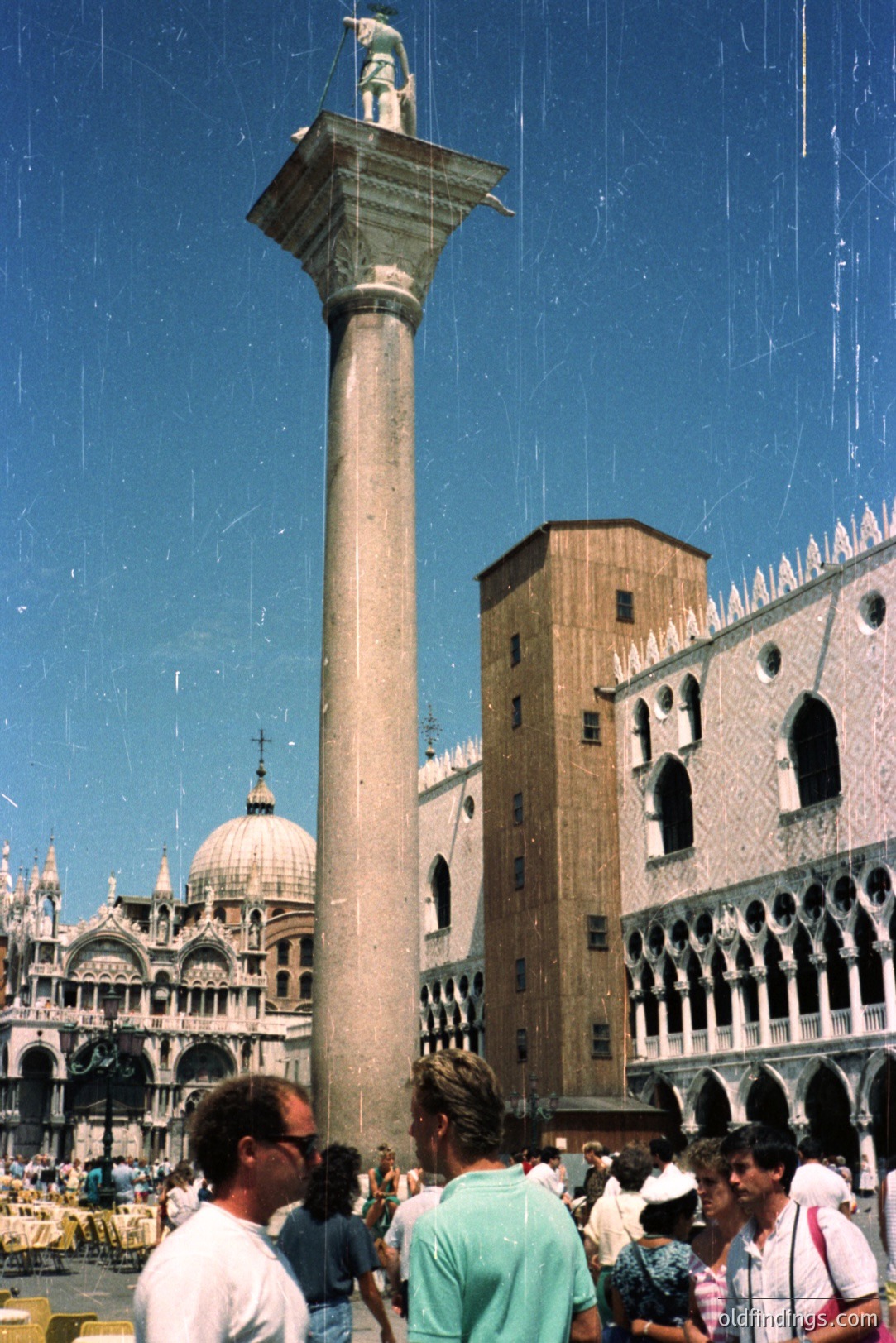 Column with statue dominates the view of Venice’s Piazza San Marco, with the Doge's Palace and St Mark’s Basilica visible in the background. Tourists are gathered at the base. Likely a 1970s travel snapshot, exhibiting characteristic film grain and color. A classic Venetian scene.