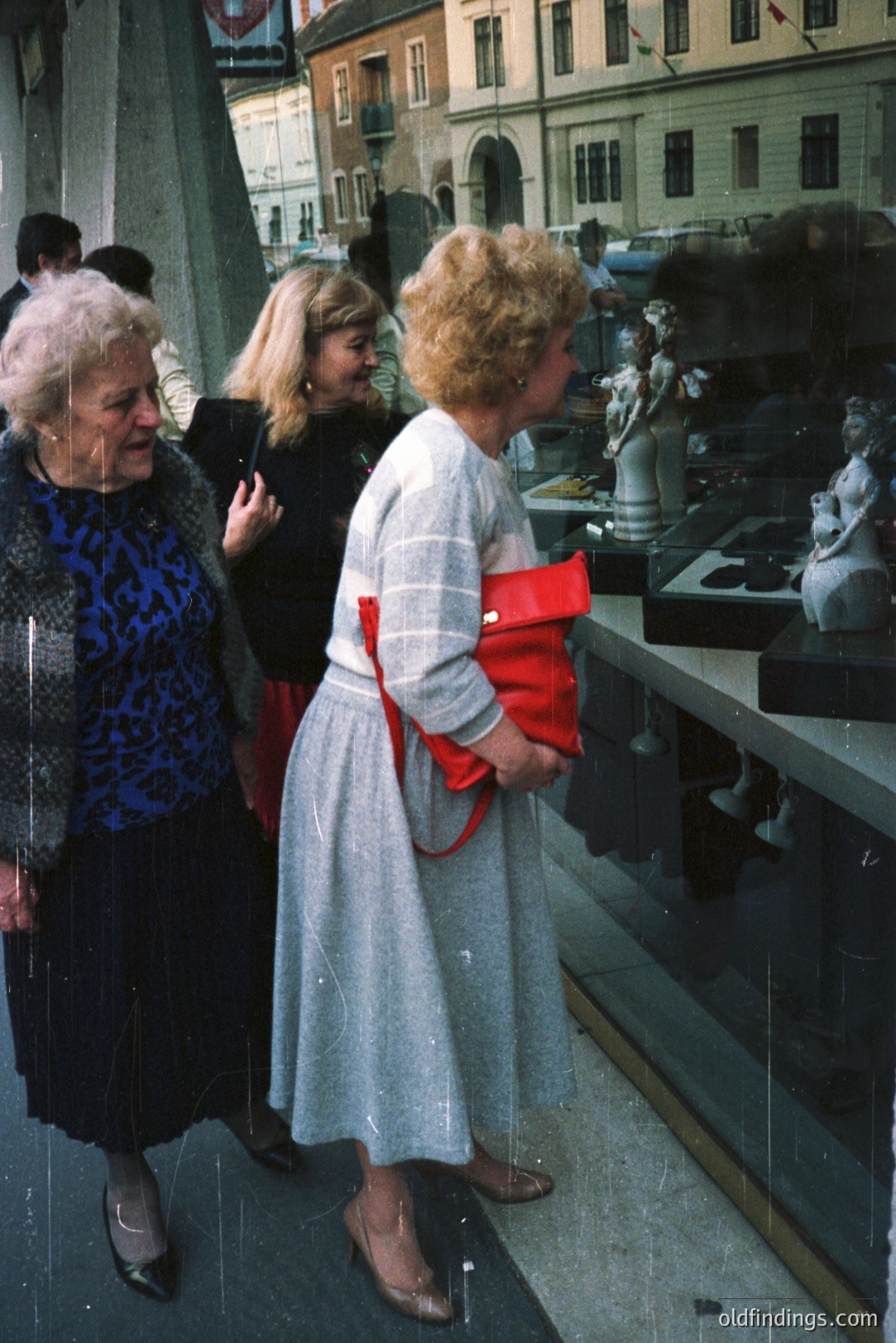 Three women examine sculptures displayed in a storefront window. The lead woman wears a blue patterned dress, the second a red jacket, and the third a gray midi dress with a red bag. Architectural details suggest a European setting. Likely 1970s fashion and style. Offers potential for vintage style design references.