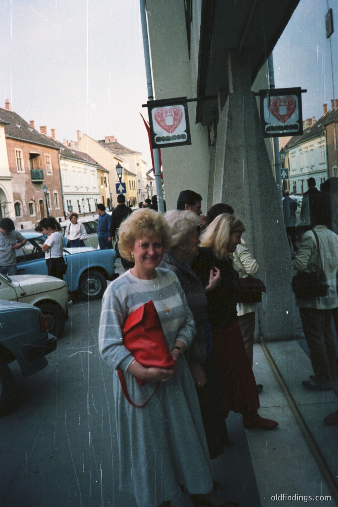 Exterior view through a shop window showing a woman in a gray dress & large hairdo holding a red purse. Visible street scene with vintage cars & pedestrians. Likely Eastern Europe, 1970s-80s, based on architecture & vehicles. Nostalgic, everyday life snapshot.