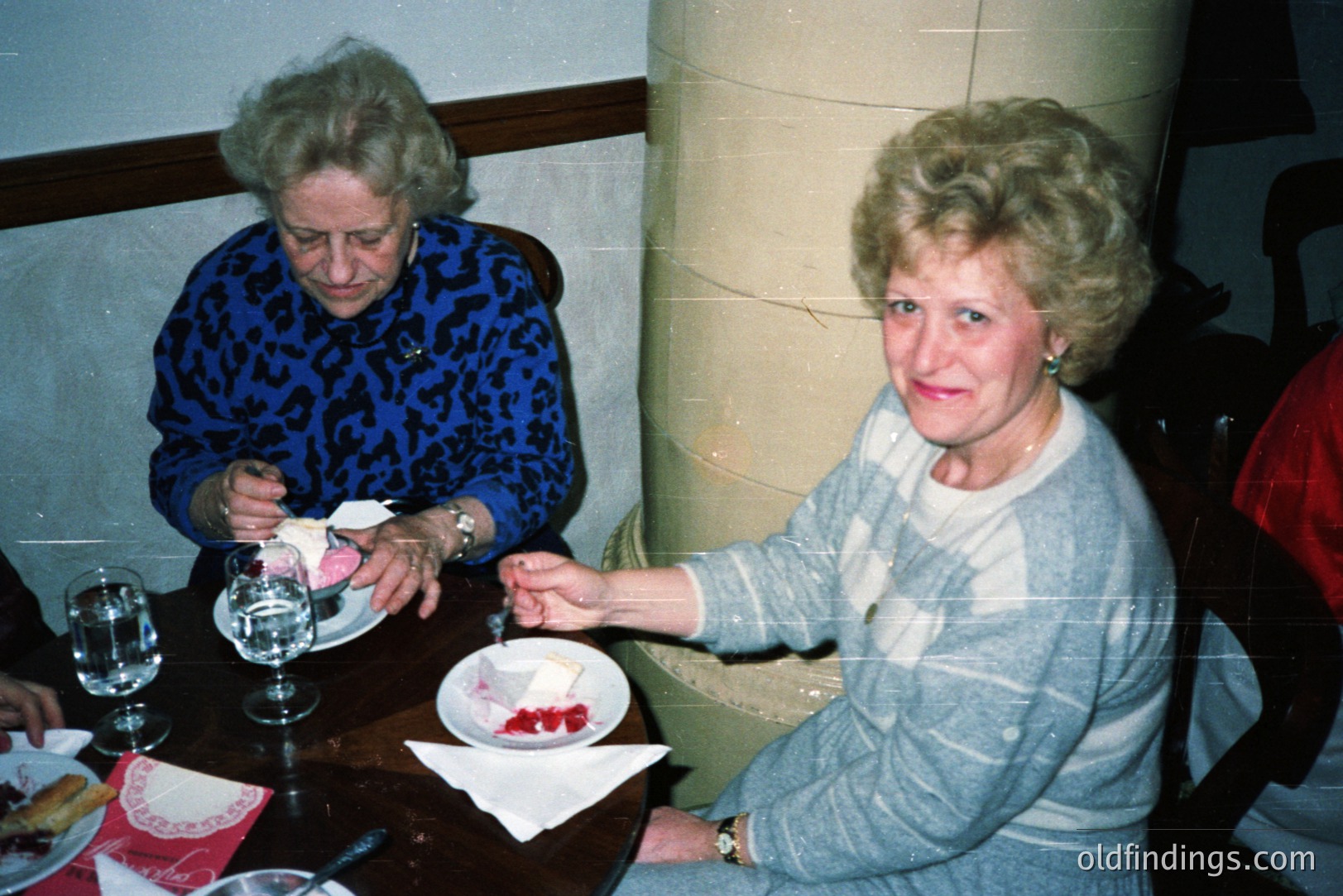 Two women seated at a table, appears to be enjoying dessert. The woman on the left wears a blue patterned sweater, while the other has a gray sweater and a white blouse. Red accents visible on table linens & background. Likely a family gathering or special occasion, perhaps 1970s-80s.