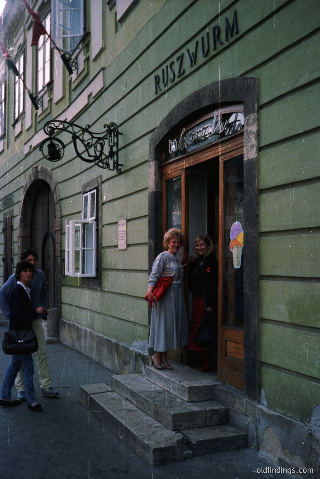 An exterior view of a building facade, displaying "RUSZWURM" signage. Two women in 1970s-era dresses stand on the entrance steps, with a third figure walking by. The architecture features a curved arch window and a decorative lamppost. Likely Eastern European, possibly Hungary.