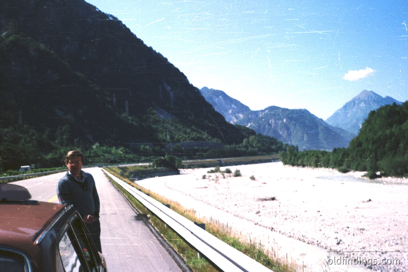 Man stands beside a vintage car on a roadside overlooking a broad, rocky riverbed framed by steep, alpine slopes and towering mountains. Likely taken in the 1970s or 80s, capturing a scenic travel moment. Vehicle and clothing suggest a leisure road trip.