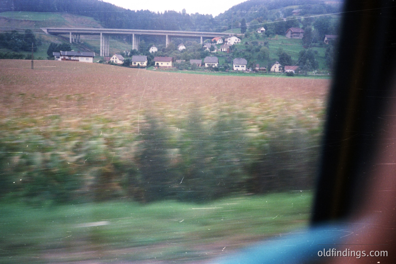 View from within a vehicle, showcasing a rural landscape with a multi-span bridge in the distance. A valley with houses and vegetation is visible, framed by forested hillsides. The image exhibits motion blur. Likely 1970s or 80s film.