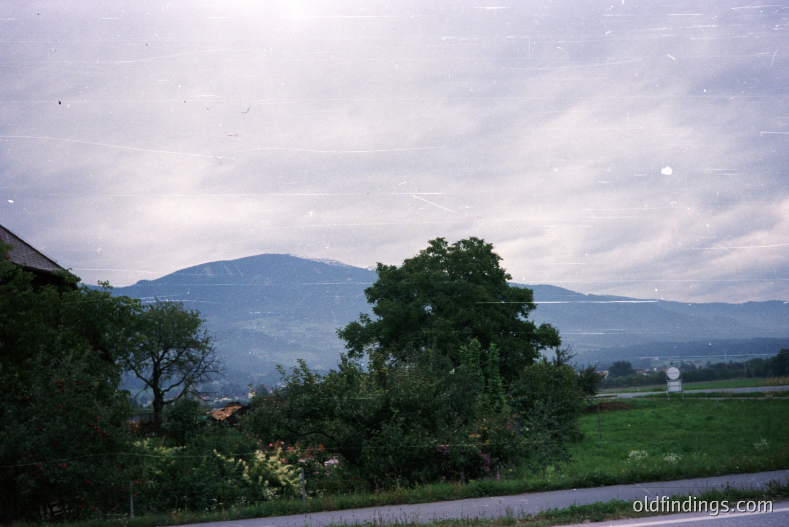 Lush, vibrant greenery dominates the foreground, framing a distant, layered mountain range under an overcast sky. A glimpse of a traditional roofline suggests a rural, European setting. Appears to be film photography. Possibly a travel or landscape stock photo.
