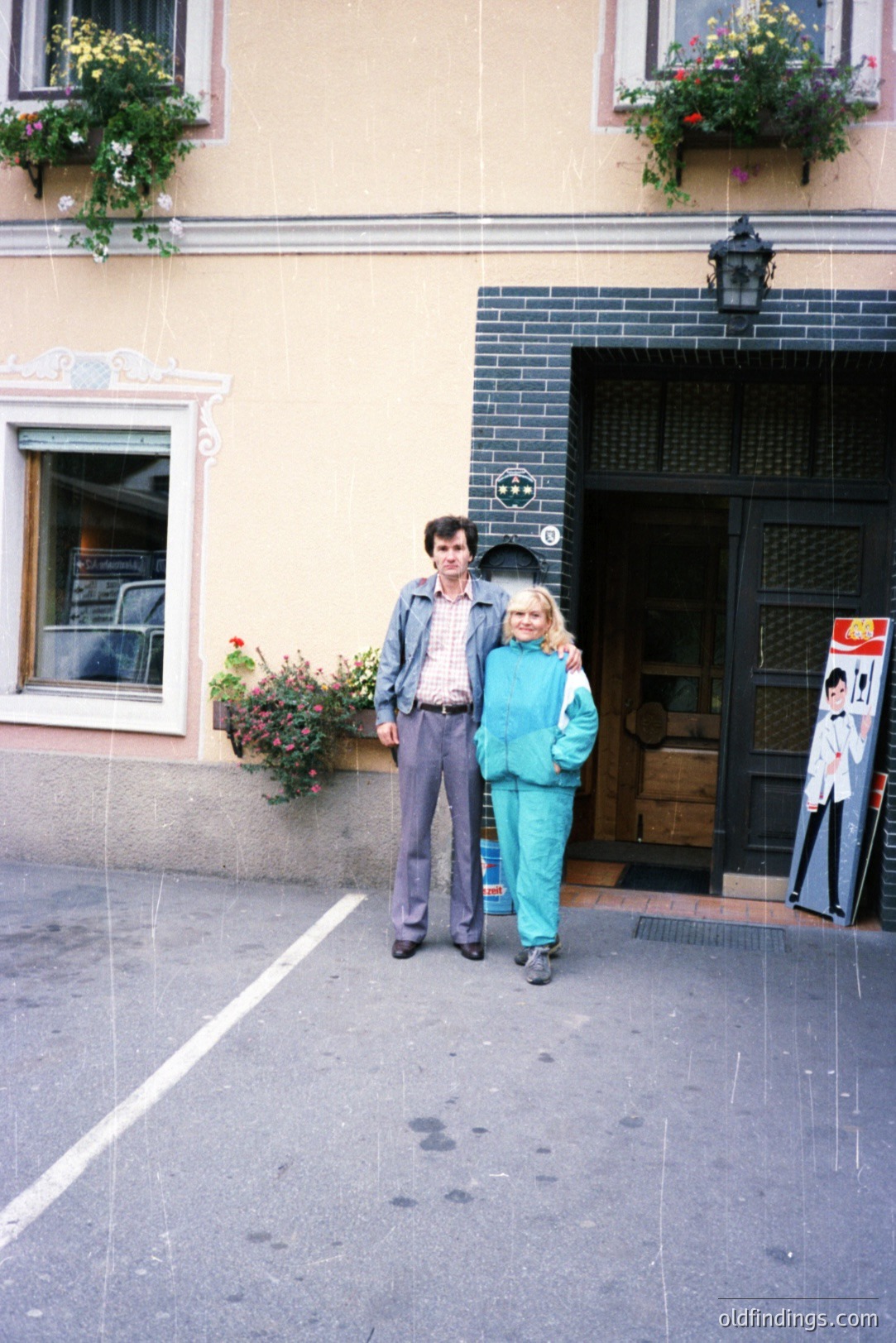 A man and woman pose outside a building with ornate window boxes and a vintage poster. The man wears a denim jacket over a shirt, and the woman a teal tracksuit. Likely 1970s style and architecture. Building details suggest a European locale. Charming street scene!