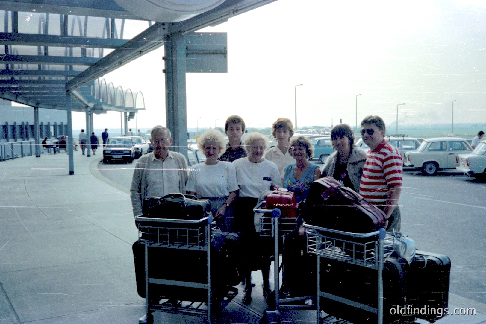 Group portrait near airport baggage claim. Six people stand with luggage carts; attire suggests a 1970s-1980s style. Cars and airport architecture visible in background. Appears to be an international travel scene. Potential for archival/family history documentation.