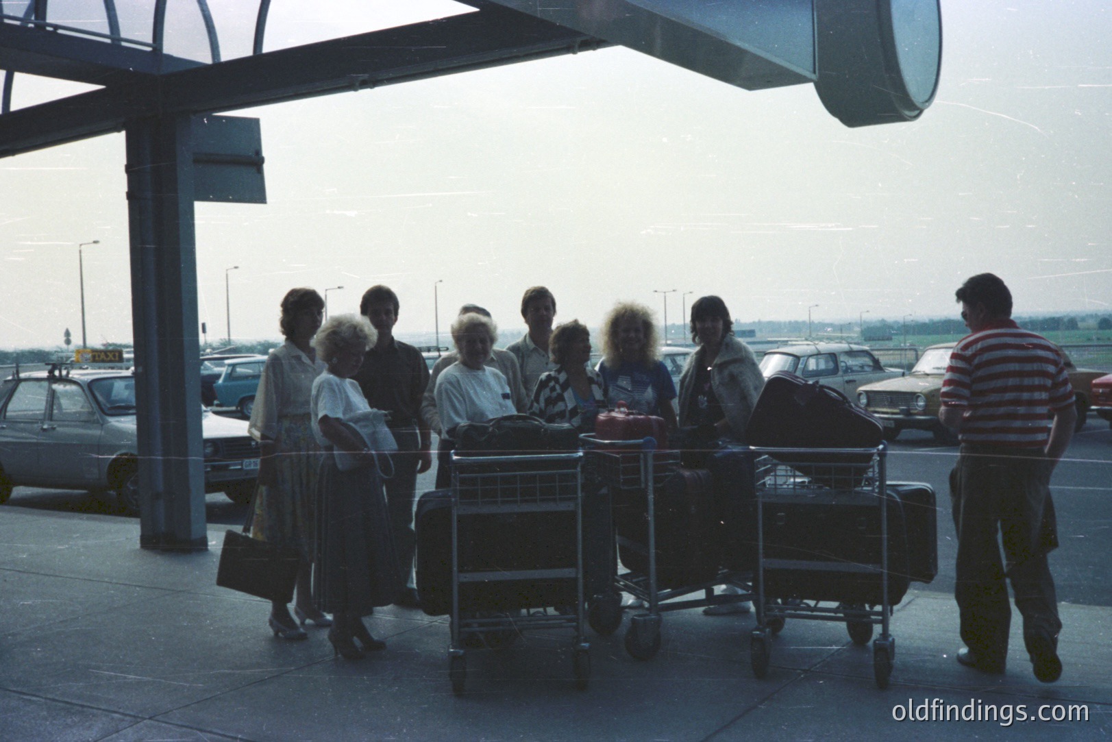 A group of travelers, likely a family, stand under a covered airport terminal. They are surrounded by stacked luggage and trolleys. Cars are visible in the background, suggesting a departure scene. Distinctive 1970s or early 1980s fashion and vehicle styles. Likely a European location. A snapshot of travel during that era.
