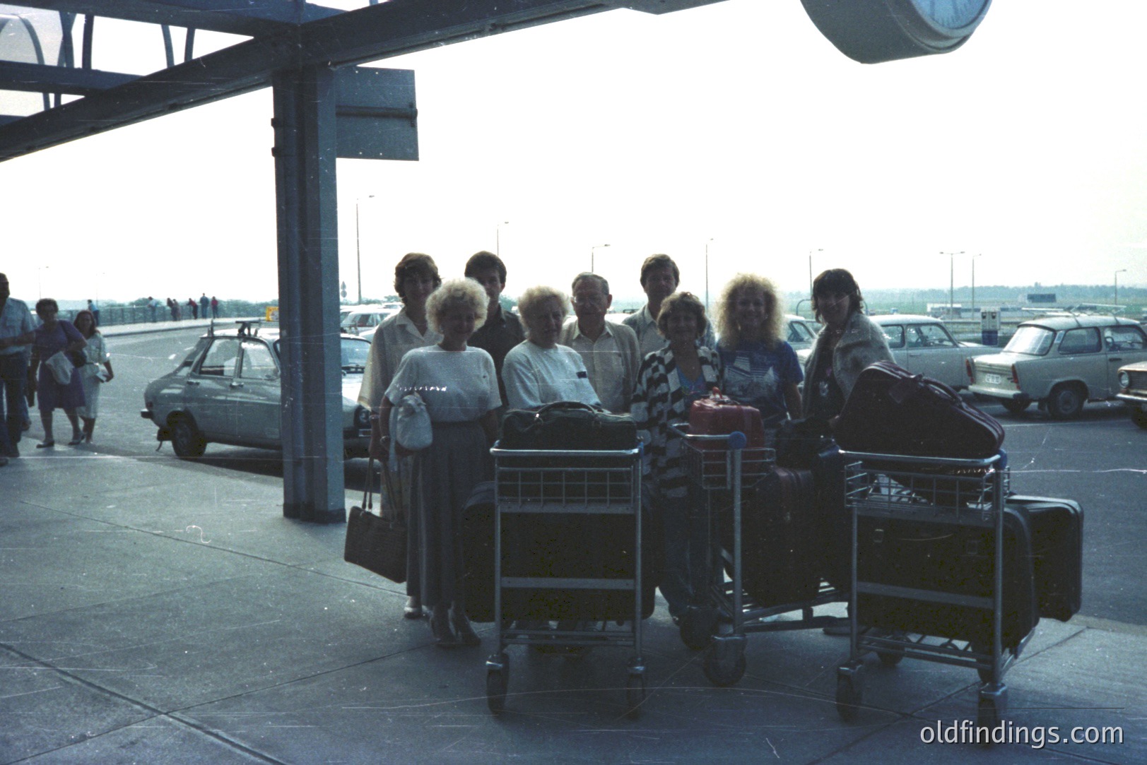 A group of eight people stand near luggage carts at an airport terminal. Visible are Soviet-era vehicles, suggesting a location in Eastern Europe. The architectural style and clothing suggest the 1970s or early 1980s. Likely a travel group or family vacation. Commercial value: design reference for vintage travel imagery.