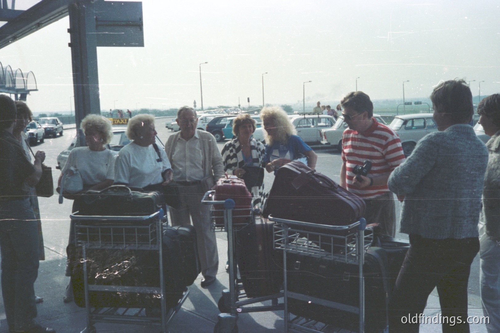 A group of vacationers, likely a family or tour group, waits at an airport curb. The scene shows luggage carts filled with bags, vintage car models, and casual 1970s/1980s attire. The photograph’s color palette & dated appearance suggests the 1970s-1980s. Potential stock photography use for travel or retro themes.