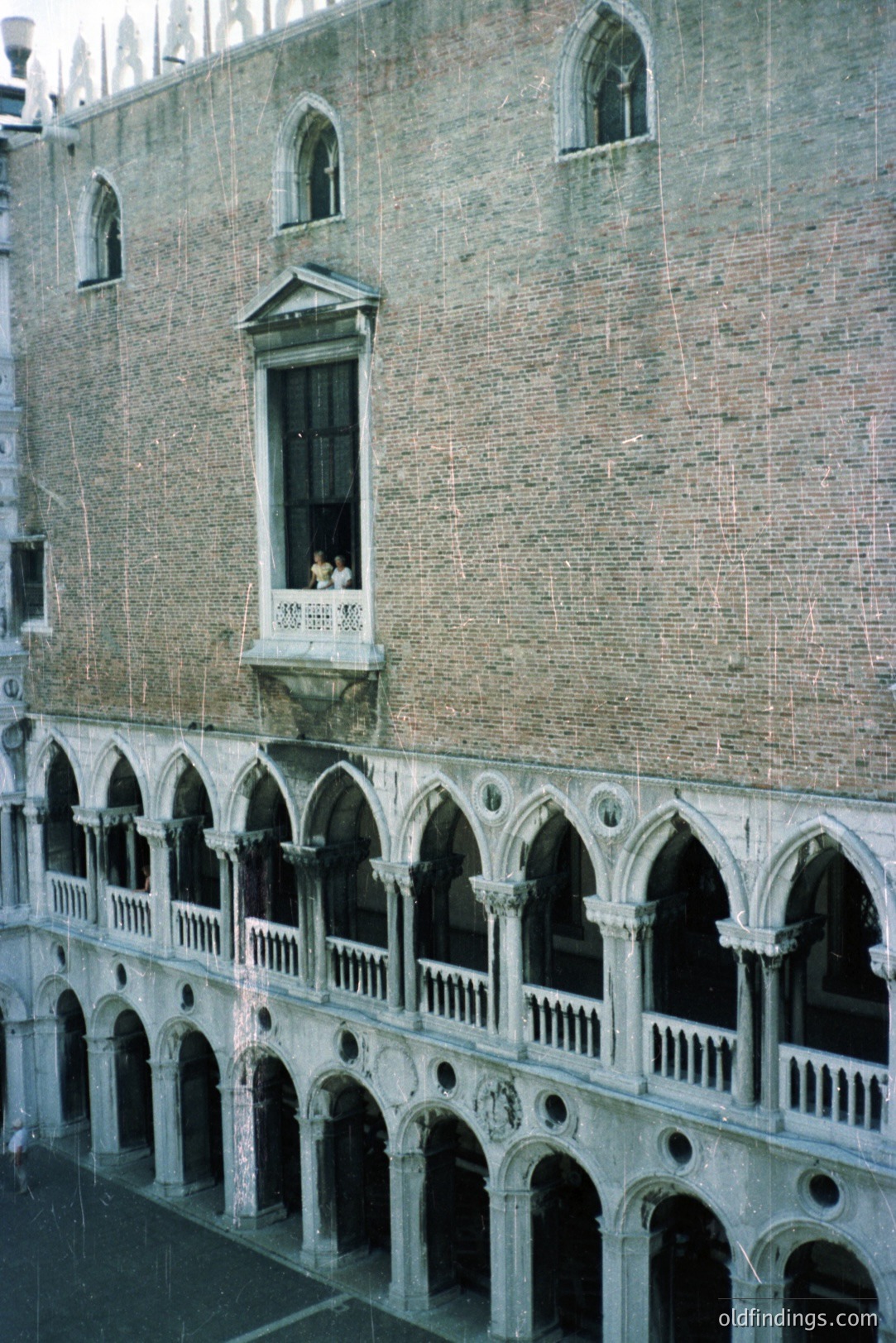 Ornate Venetian palazzo facade showcases arches and a balcony with figures. The building's brickwork displays intricate detailing characteristic of Venetian Gothic architecture. Likely photographed in the 1970s, capturing a historic urban scene. Potential use for architectural stock or design references.