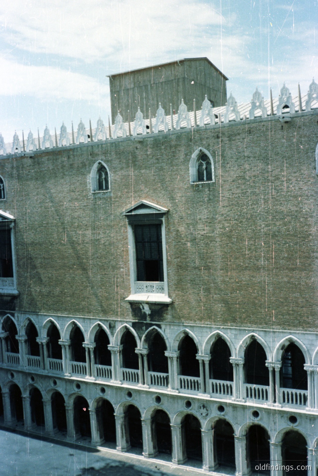 Grand canal façade of the Doge’s Palace in Venice, Italy. Features ornate Gothic tracery on the lower loggia, contrasting with a plain brick upper facade and decorative battlements. A chimney is prominently visible. Likely taken mid-20th century. Architectural reference for design.