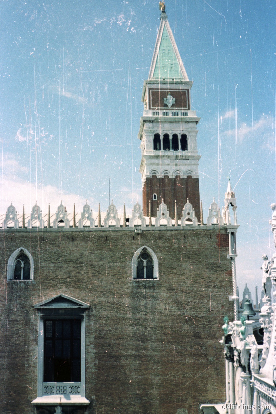 Okay, here's a description of the photograph, formatted as requested: Close-up of a brick facade featuring arched windows and a distinctive, tiered bell tower. The tower's upper levels have small, rectangular windows, topped with a green spire. Likely Venice, Italy. The aged photo shows slight discoloration and image noise.