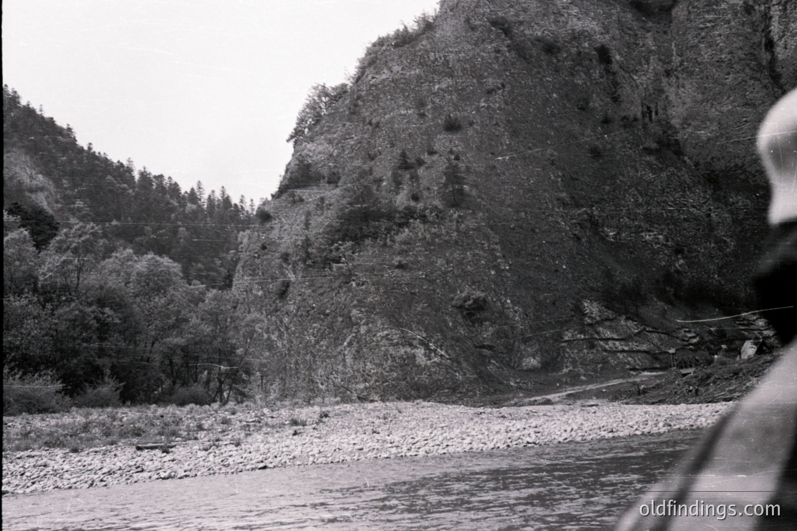 A rocky riverbed flows toward a steep, forested hillside and a dramatic rock face. A narrow road clings to the cliffside. Partial view of a person's shoulder and plaid clothing in foreground. Possibly a scenic overlook or hiking trail. Likely 1970s landscape photography.