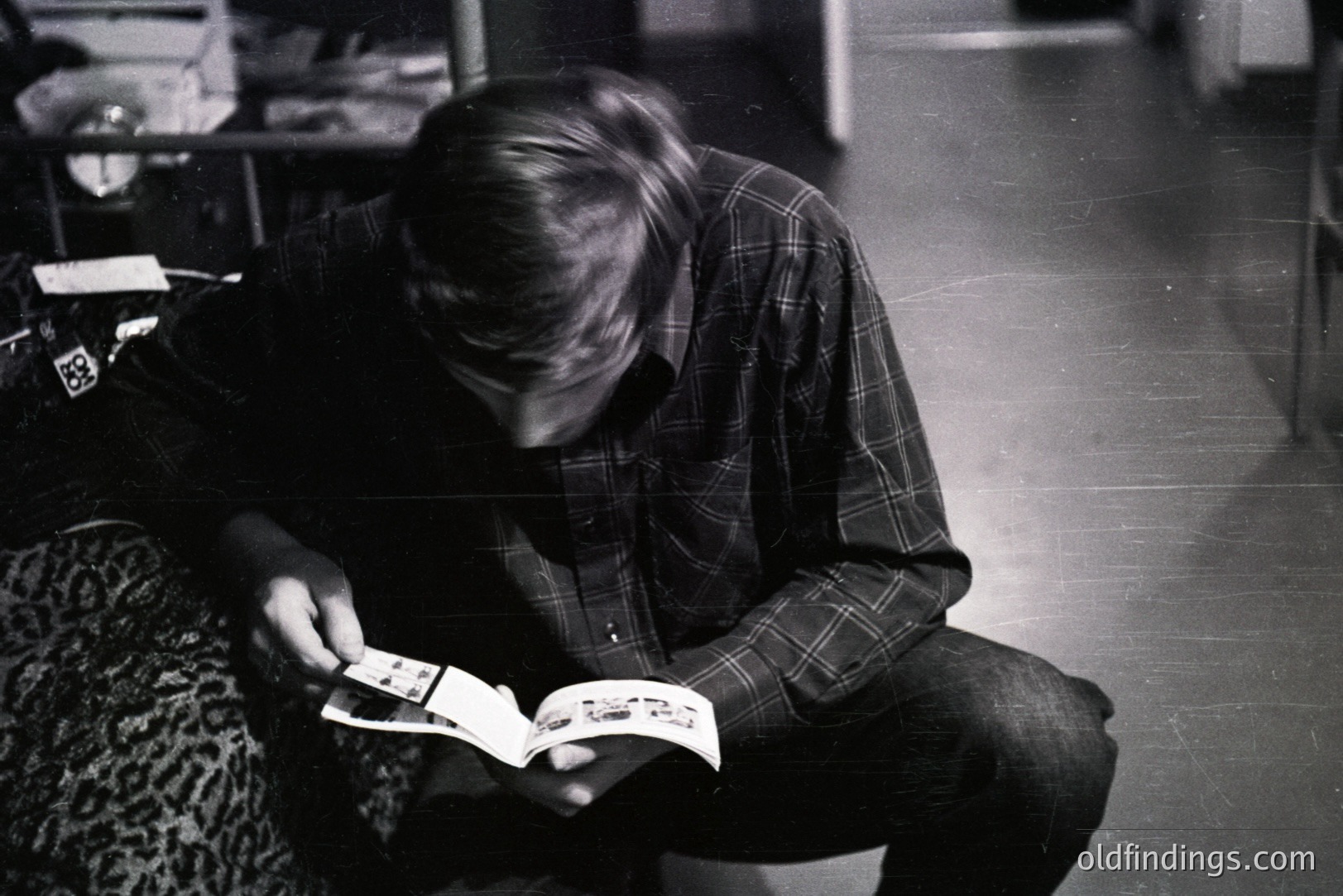 A young man, seated on a patterned sofa, intently examines a booklet or magazine. He wears a plaid shirt and dark trousers. The scene suggests a casual, domestic setting; possibly 1970s. A glimpse of printed material within the booklet hints at a commercial or informational context.
