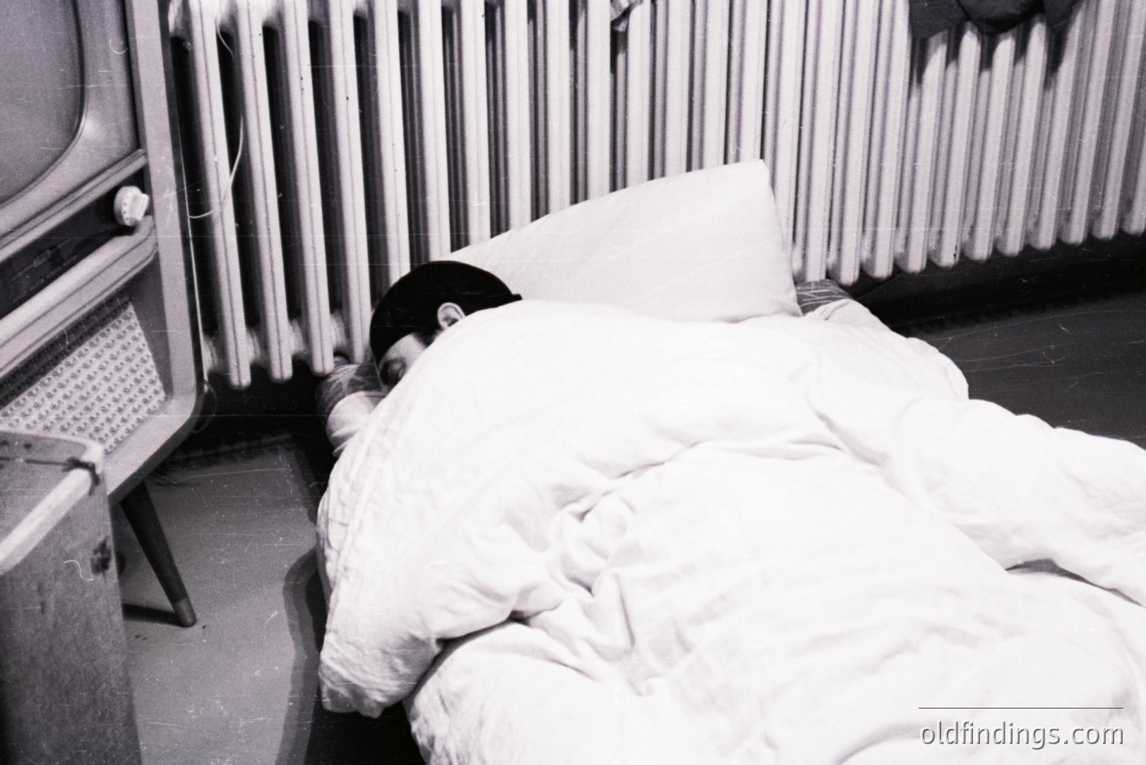 A figure lies beneath a heavy duvet, partially obscured, with only a hand visible. A vintage television set and radiator are visible in the background, suggesting a domestic interior. Appears to be a candid moment, likely 1970s style. Suitable for stock photography relating to sleep, privacy or retro design.