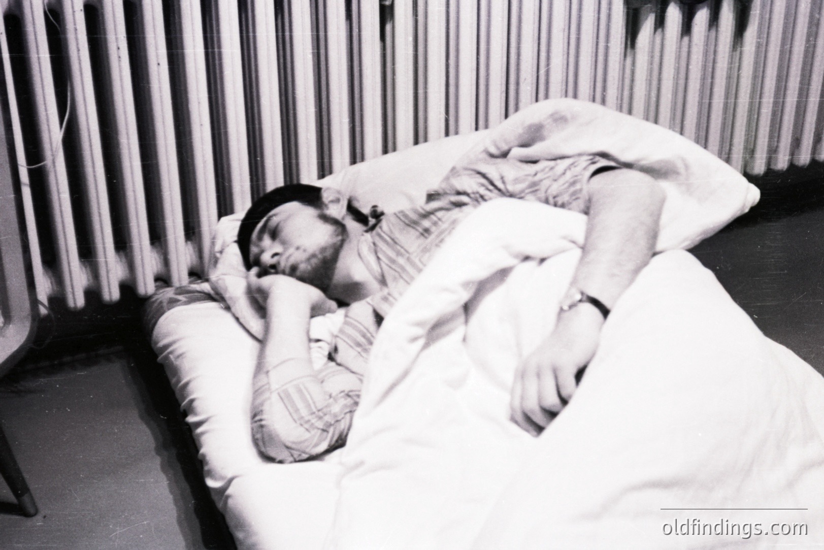 Man asleep in bed, covered with a blanket and wearing a dark cap. Visible wristwatch on his arm. Minimalist setting with a radiator visible in background. Appears to be a candid, potentially documentary-style image. Likely 1970s-1980s, based on aesthetic.