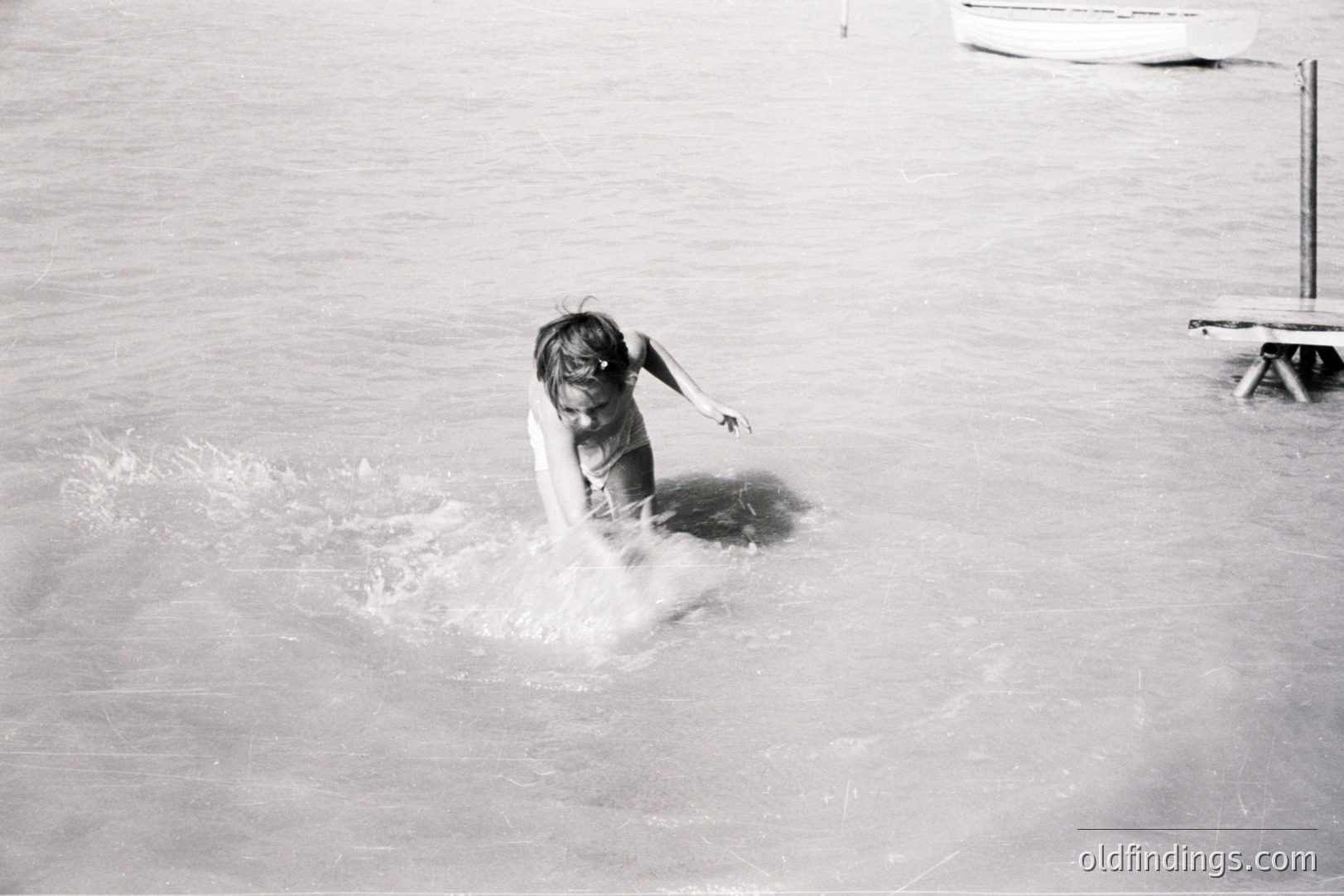 A young girl playfully splashes into shallow water. Visible details include a simple wooden dock and a portion of a boat in the background, suggesting a seaside location. The image’s grainy quality indicates a vintage film photograph, possibly from the 1960s or 70s. Captures a candid moment of childhood recreation.